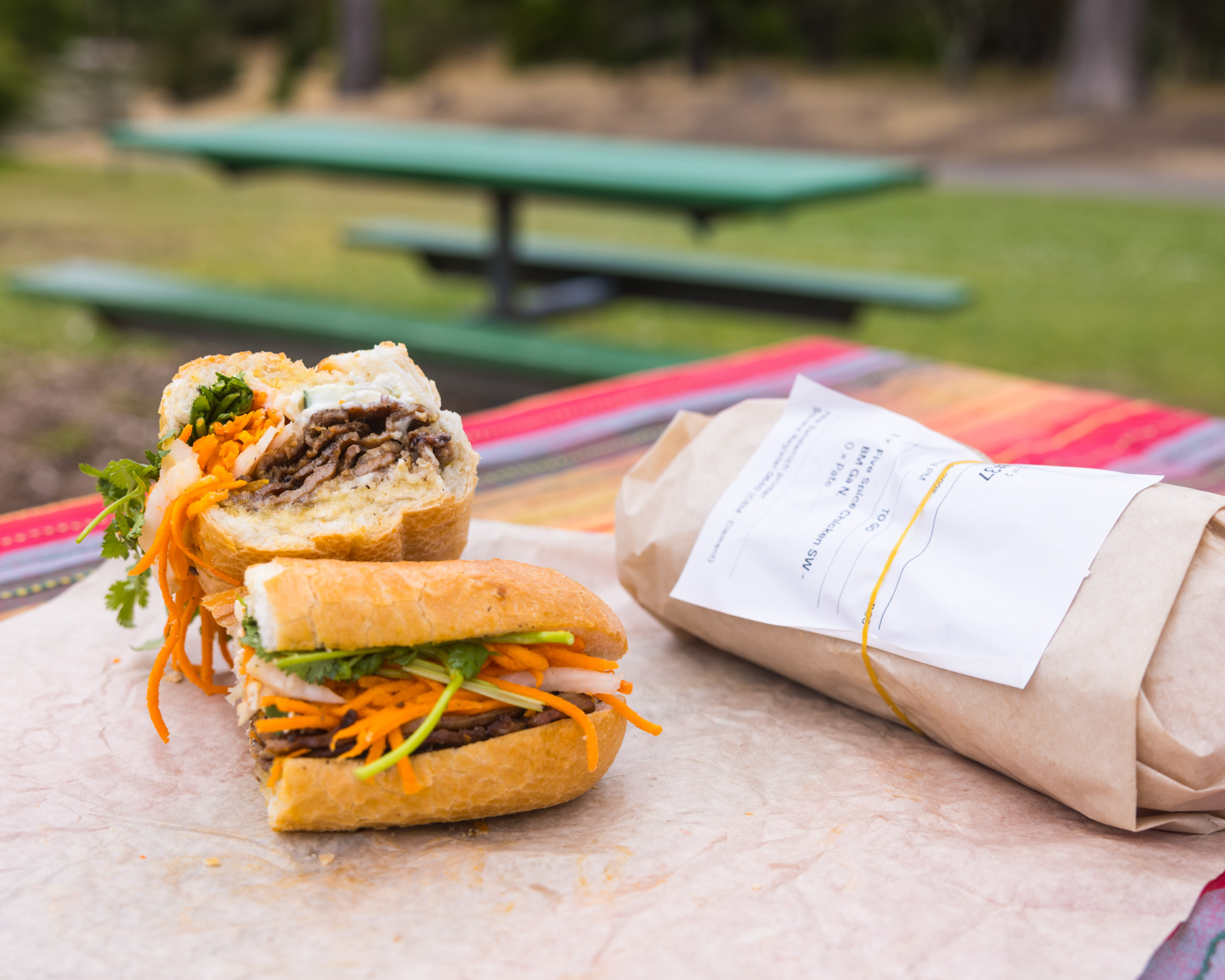 A sandwich filled with sliced meat, carrots, and herbs is on a table with colorful cloth. Next to it is a wrapped package, possibly another sandwich.