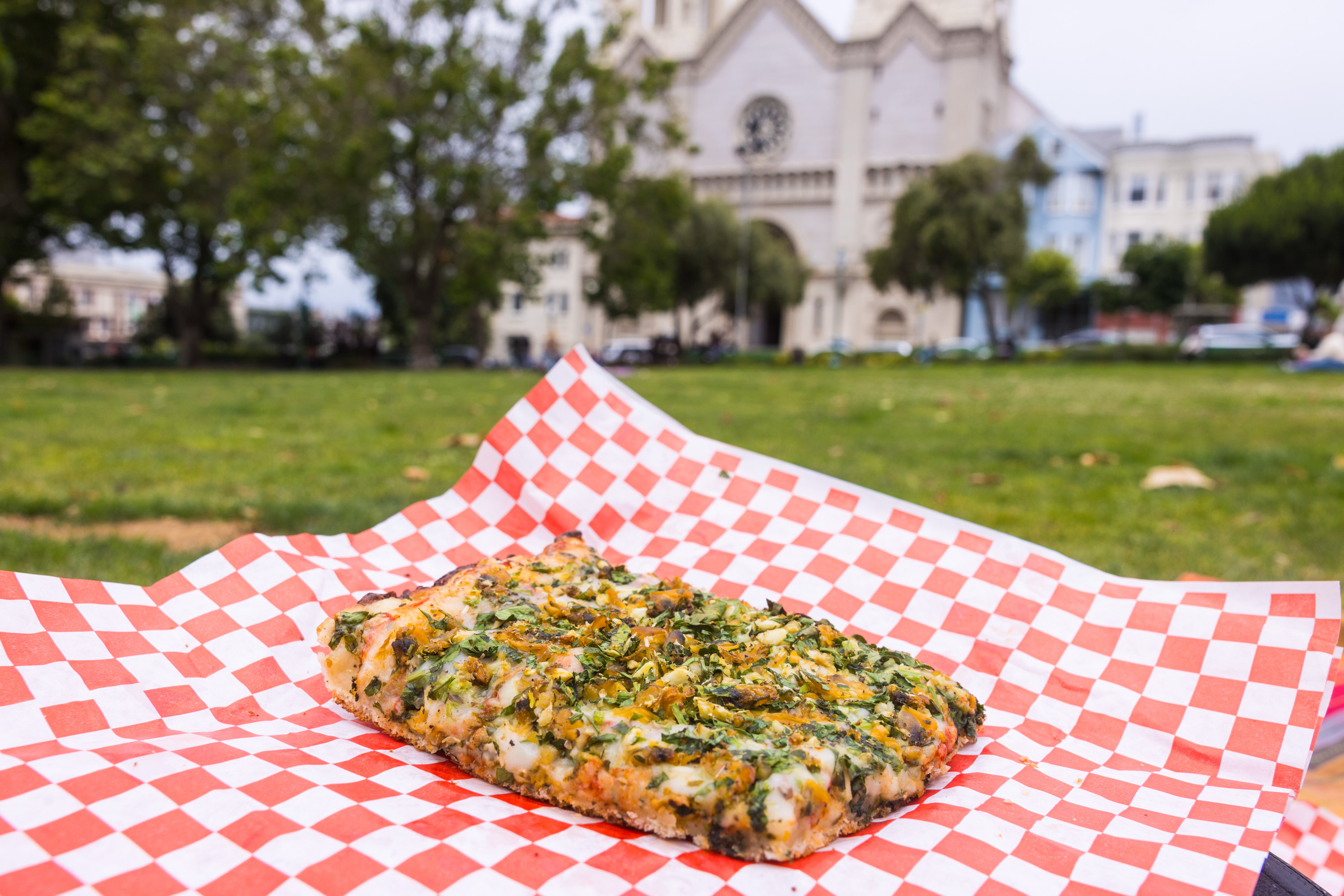 A slice of pizza with herbs is on red-checkered paper, set on grass. In the background, there's a church and trees.