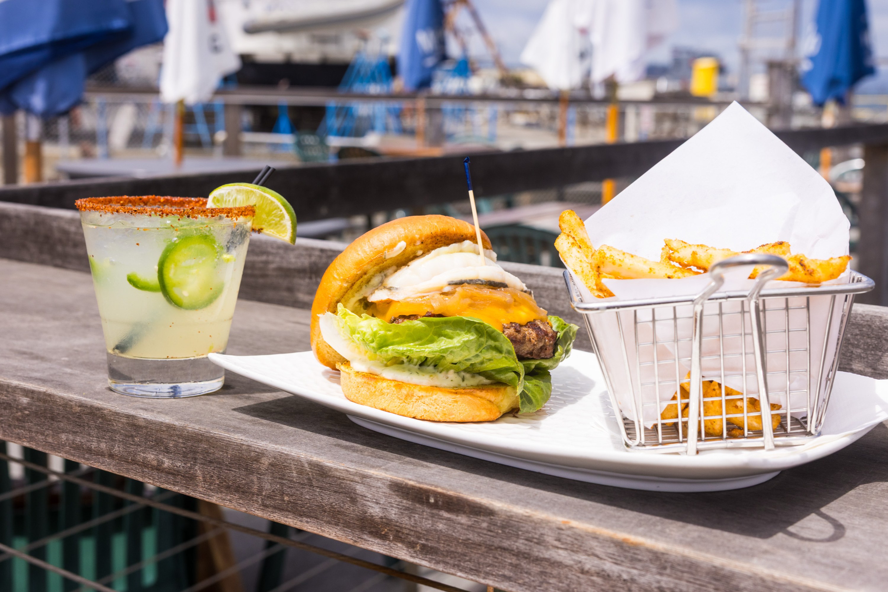 A cheeseburger with lettuce and onion sits on a plate with a basket of fries. A cocktail garnished with lime and jalapeño is beside it; a boat and umbrellas appear in the background.