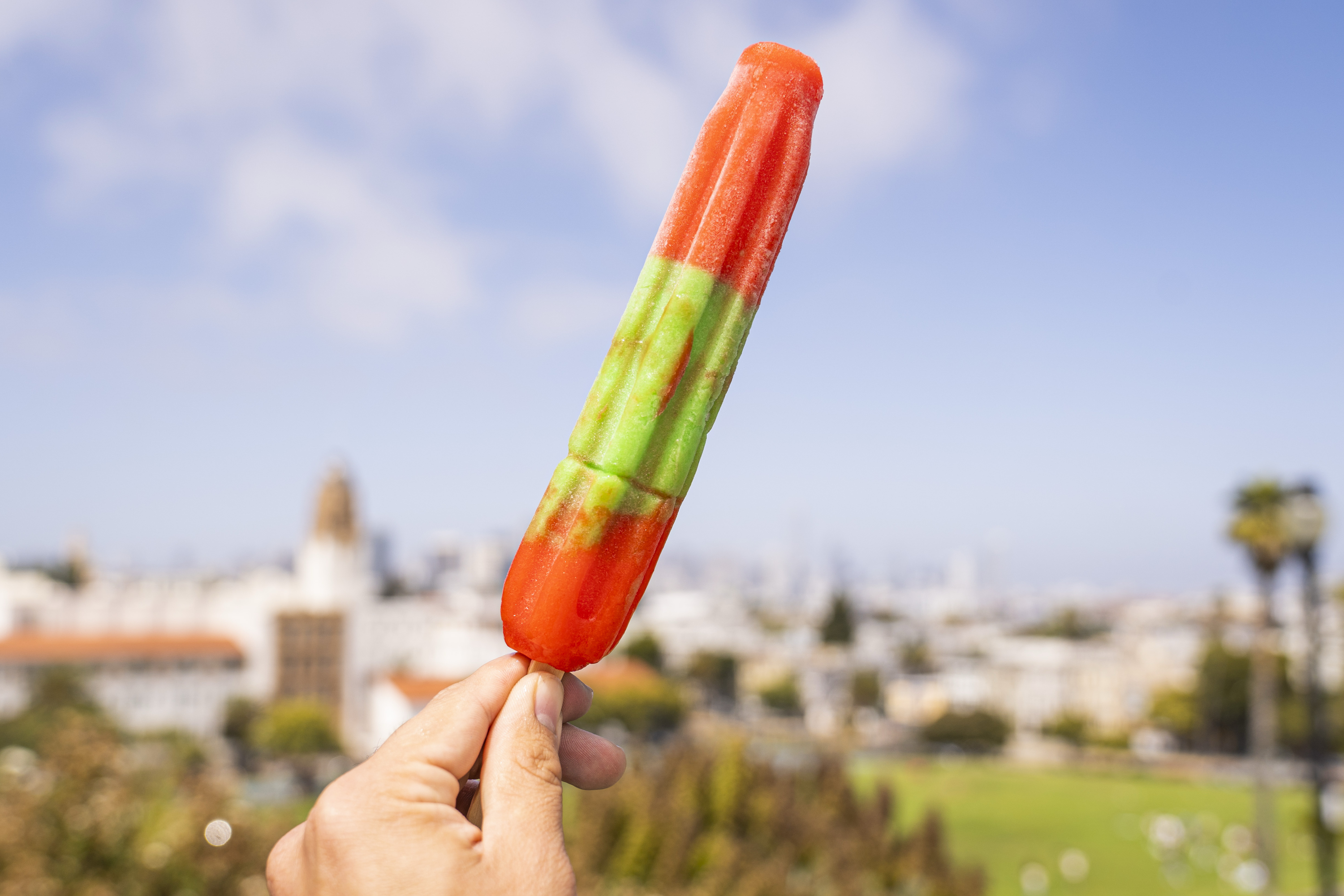 A hand holds a colorful popsicle with red, green, and orange sections against a blurred cityscape backdrop and blue sky.