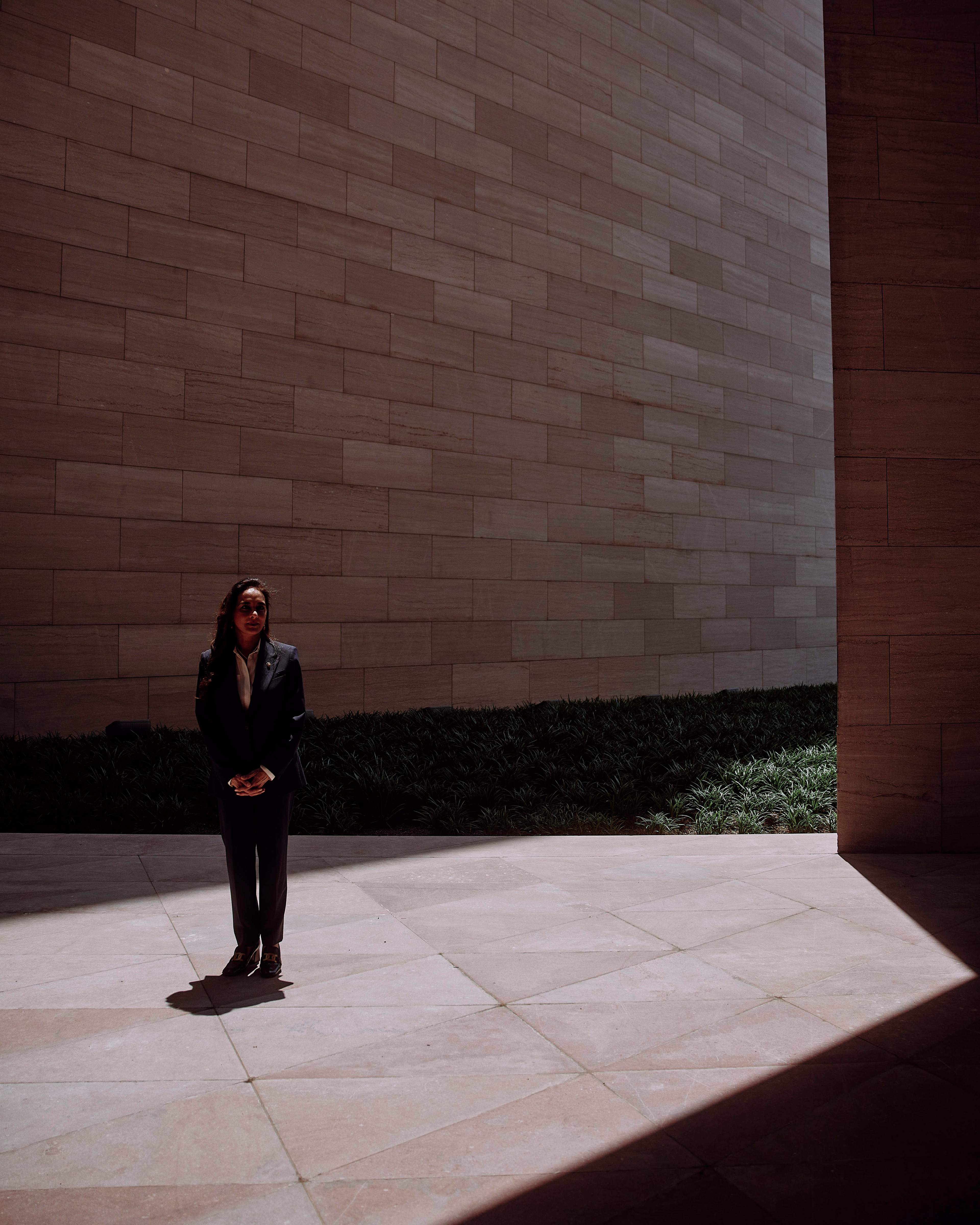 A person in a dark suit stands in a sunlit courtyard, surrounded by large tiled walls and patches of greenery in the background.