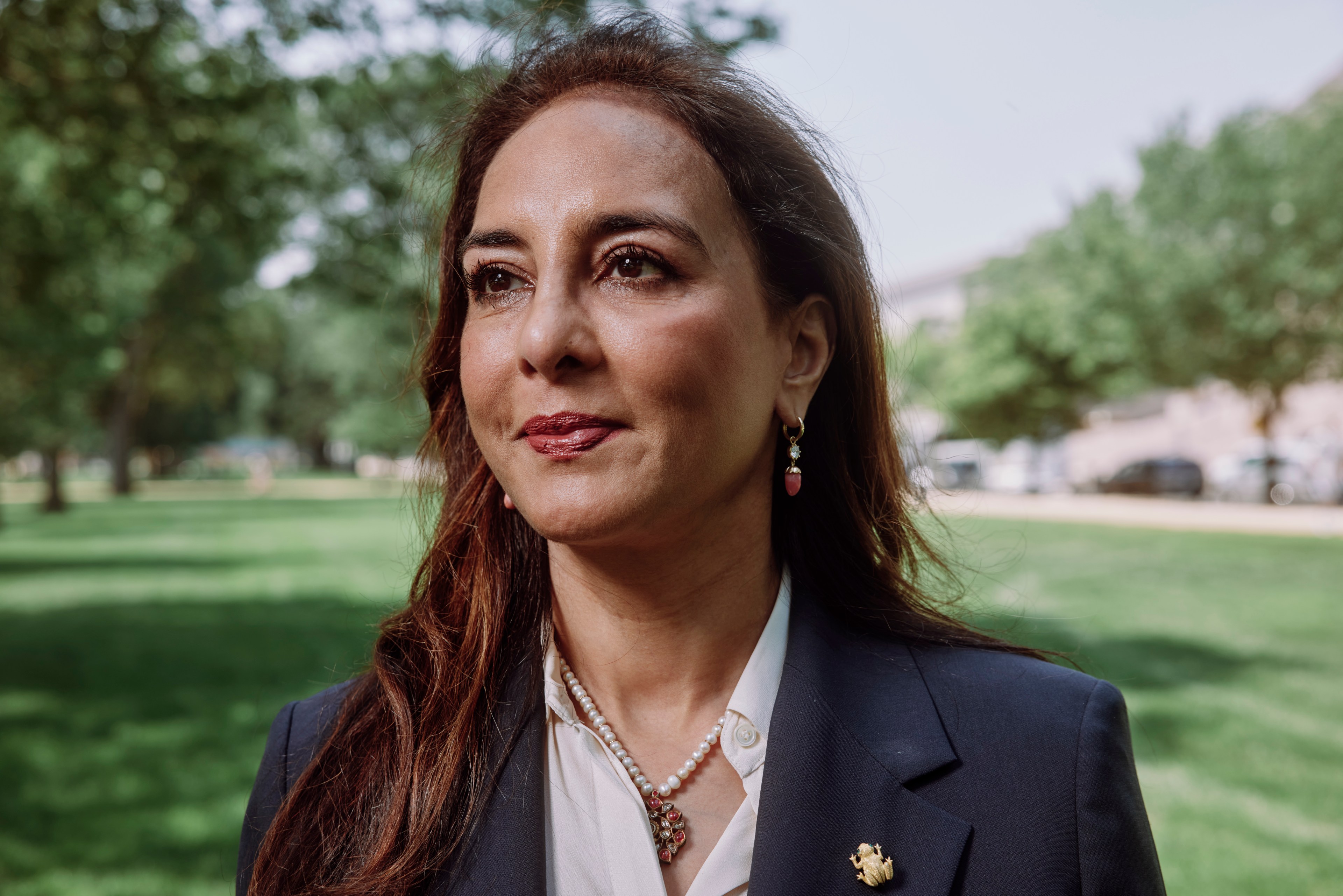 A woman with long brown hair, wearing earrings and a navy blazer, stands outdoors in a sunny park with green grass and trees in the background.