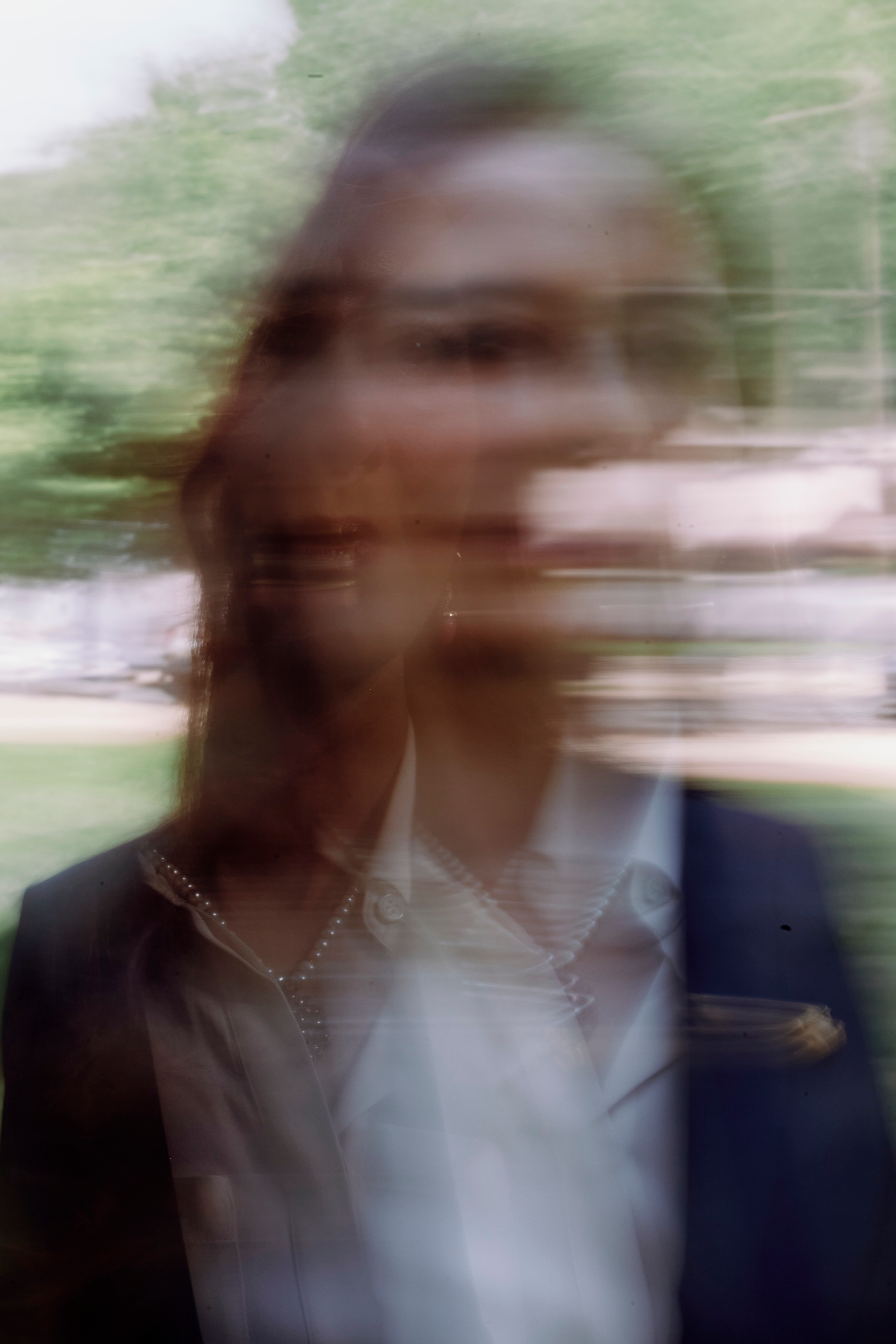 The image shows a blurred, double-exposed portrait of a person outdoors, with greenery in the background, giving a ghostly, distorted effect.