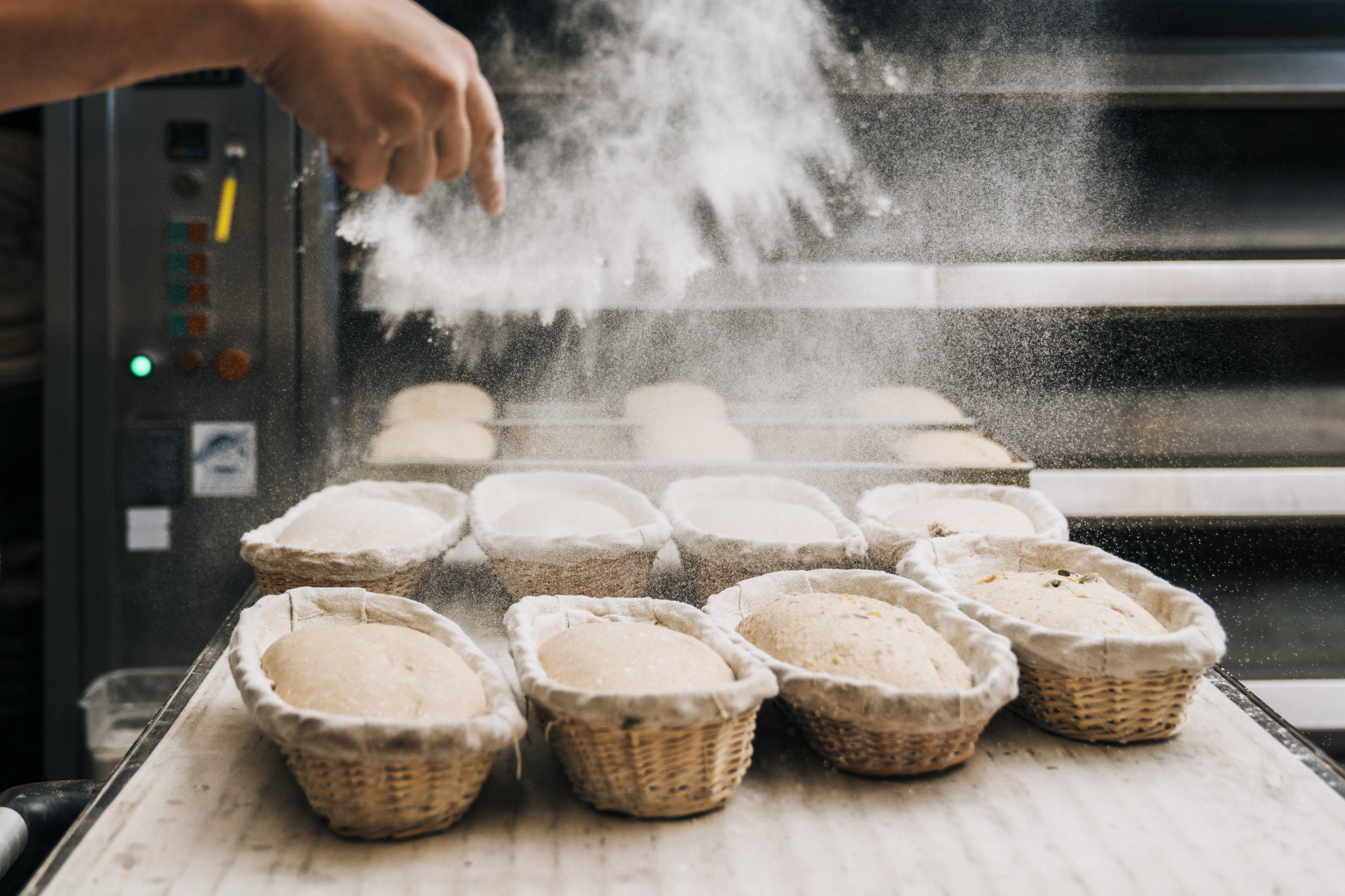 A hand sprinkles flour over dough loaves in wicker baskets, set on a table with a baking oven in the background.