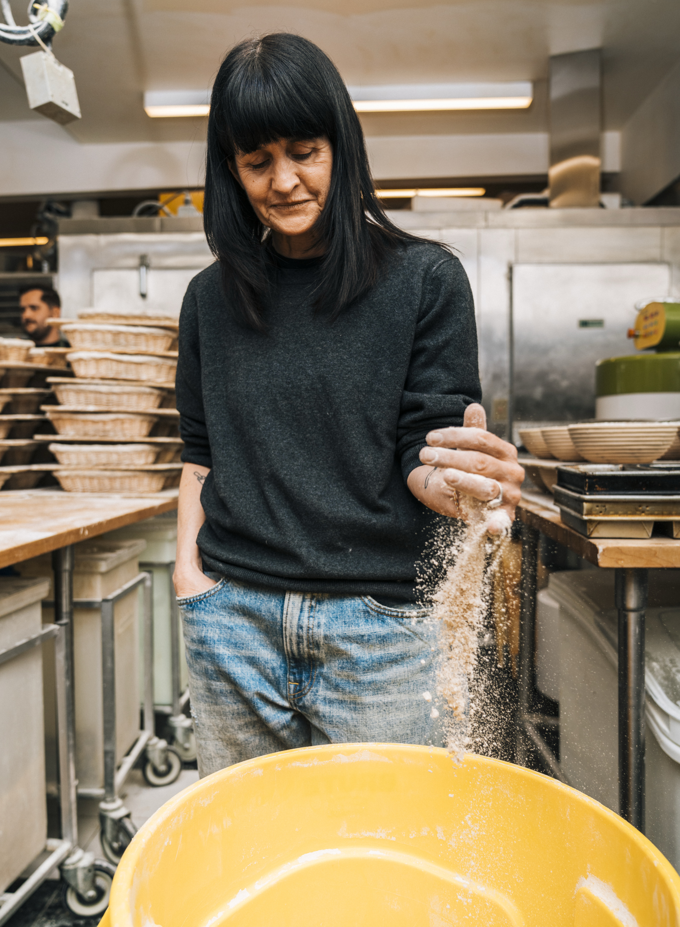 A person in a kitchen sprinkles flour into a yellow container, wearing a dark sweater and jeans. Stacks of baskets are visible in the background.
