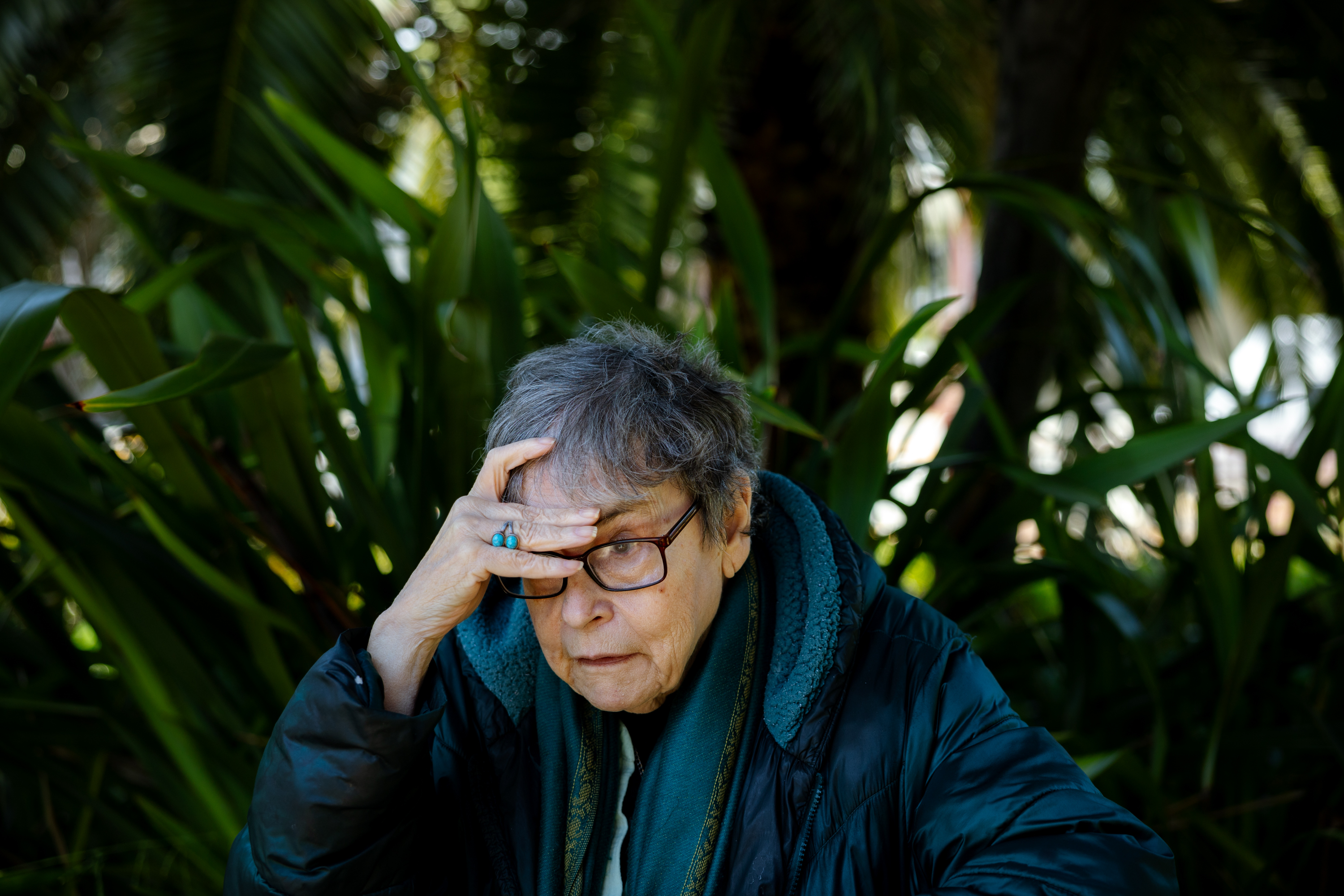 An older person with glasses looks thoughtful while resting their hand on their forehead. They sit outdoors surrounded by large green leaves.