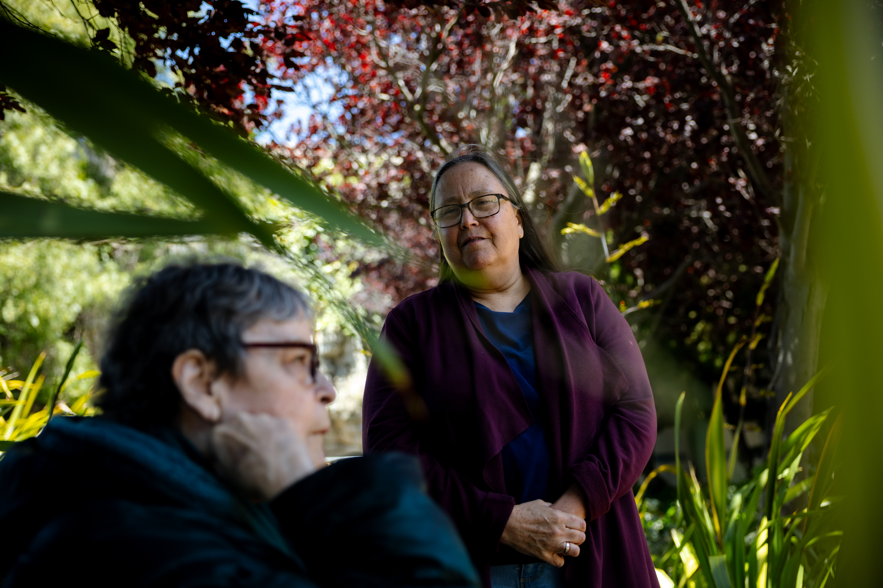 Two people stand outdoors among greenery. One is in focus, wearing glasses and a purple cardigan, while the other is blurred in the foreground.
