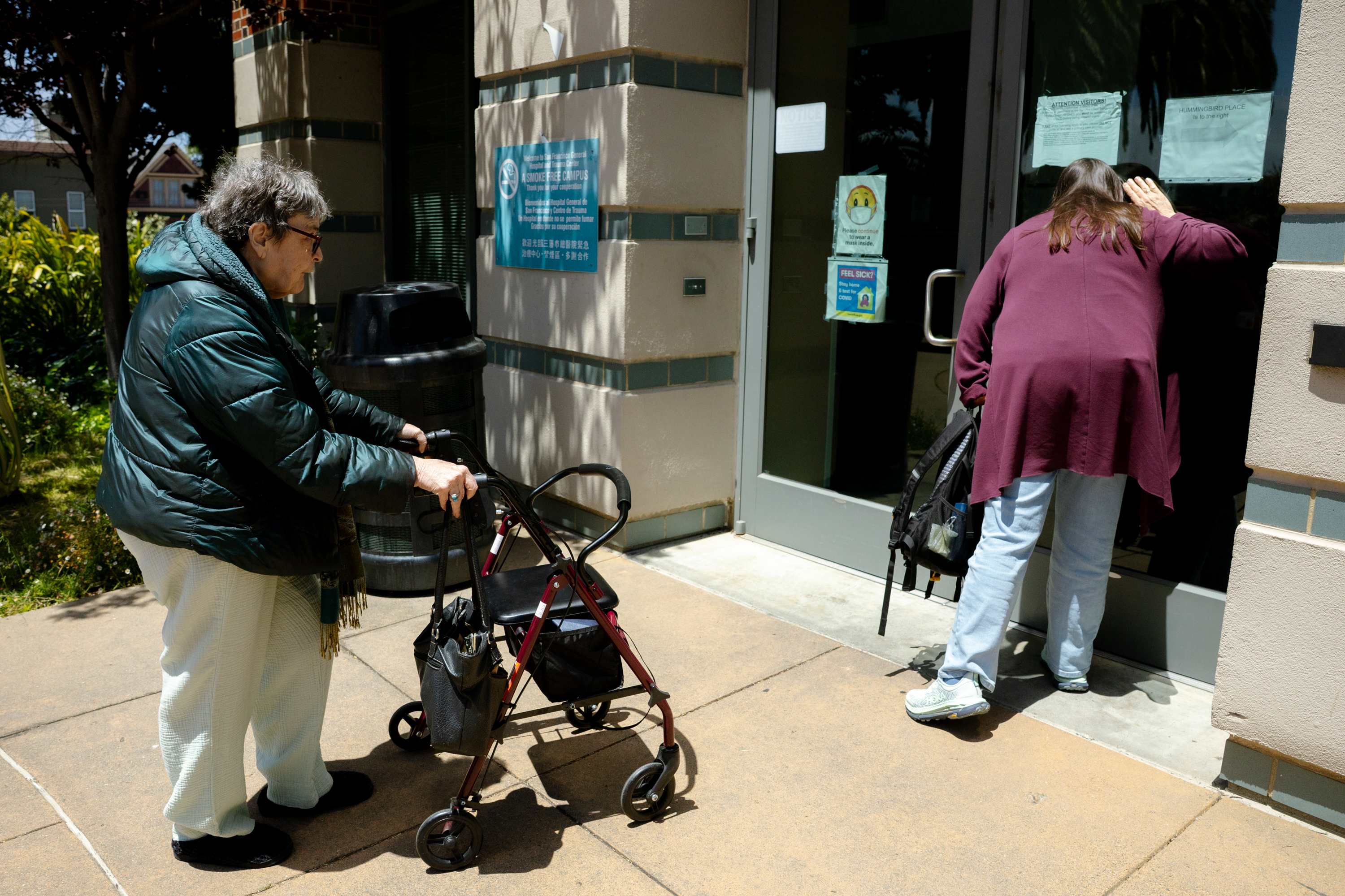 Two people are outside a building. One uses a walker, and the other leans against a glass door, looking inside. Signs are visible on the wall and door.