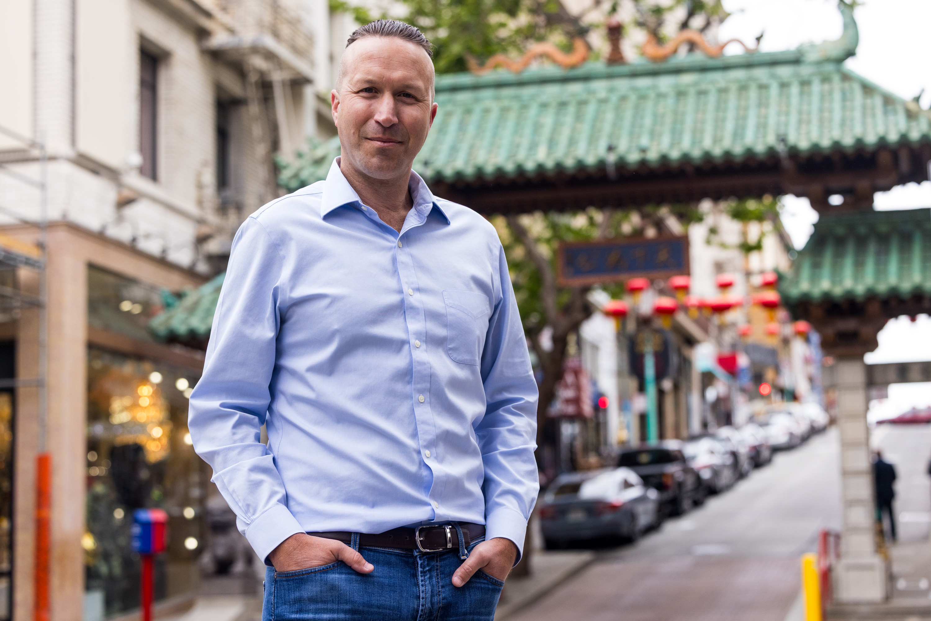 A person in a light blue shirt and jeans stands with hands in pockets on a street with cars lined up, near a traditional green-roofed archway.