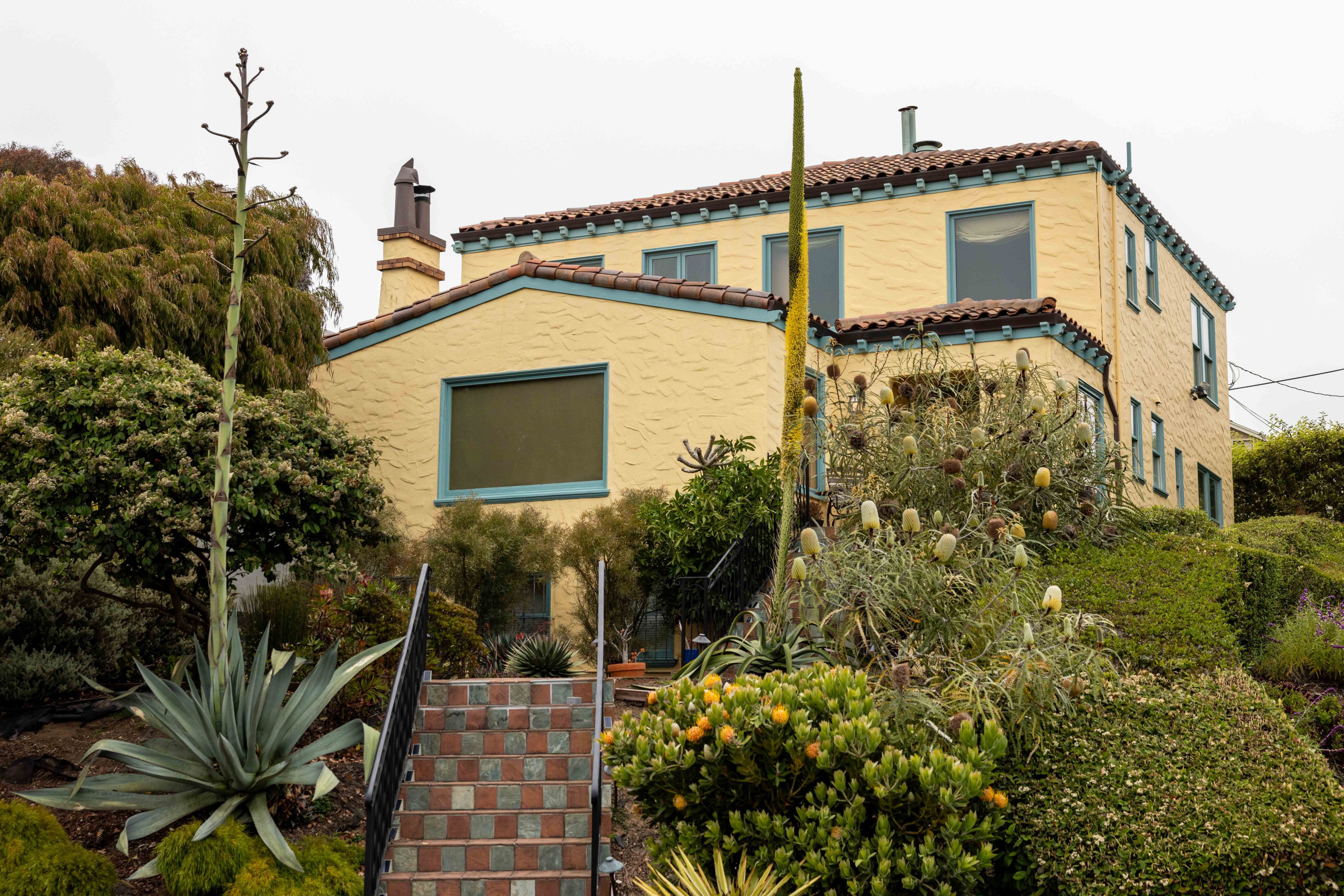 A yellow, two-story house with blue window frames and a red-tiled roof is surrounded by lush greenery and desert plants, with tiled steps leading up to it.