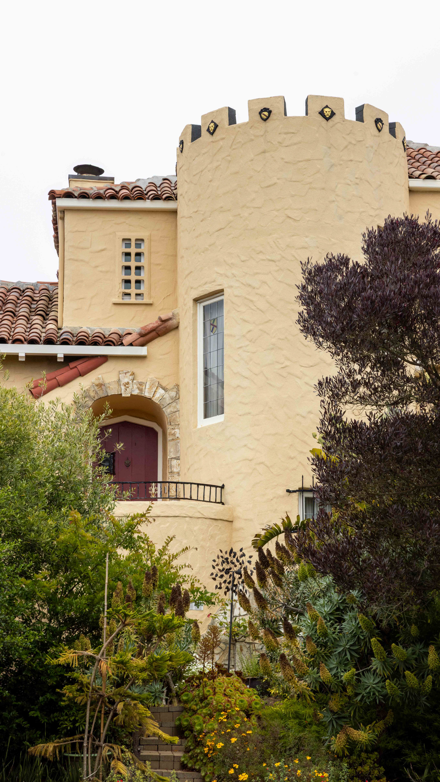 A beige stucco house with a tower-like structure and red tiled roof is partially visible, surrounded by lush greenery and tall plants.
