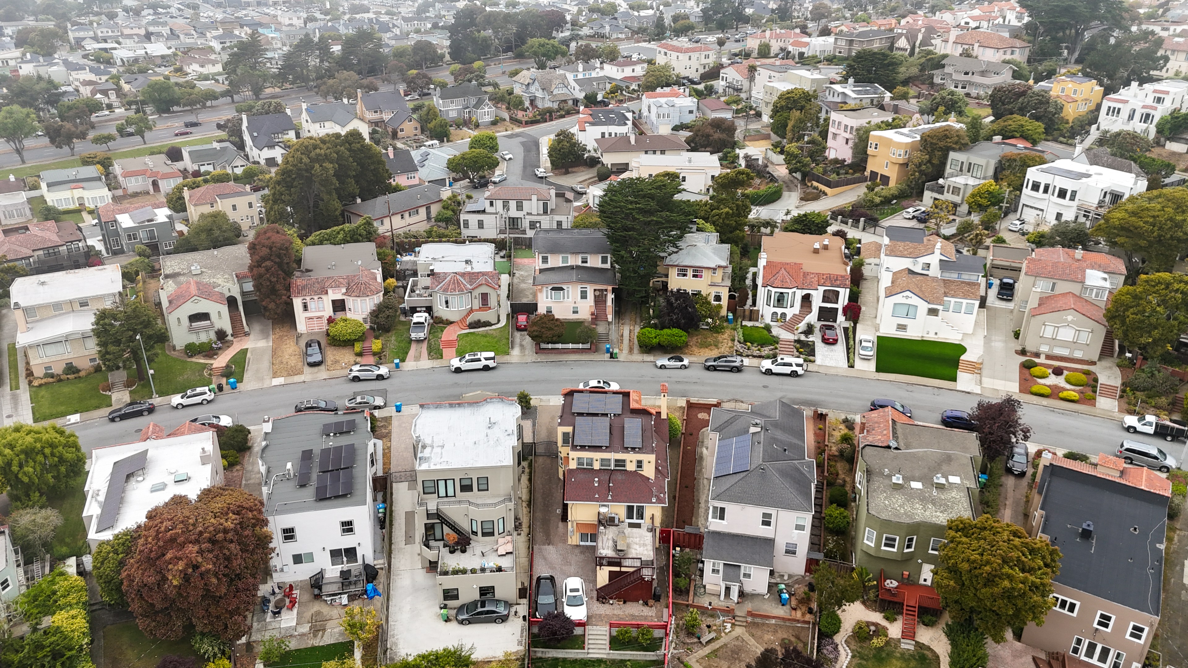 Aerial view of a residential neighborhood featuring houses with varied architecture, lush trees, and parked cars lining the curved streets.