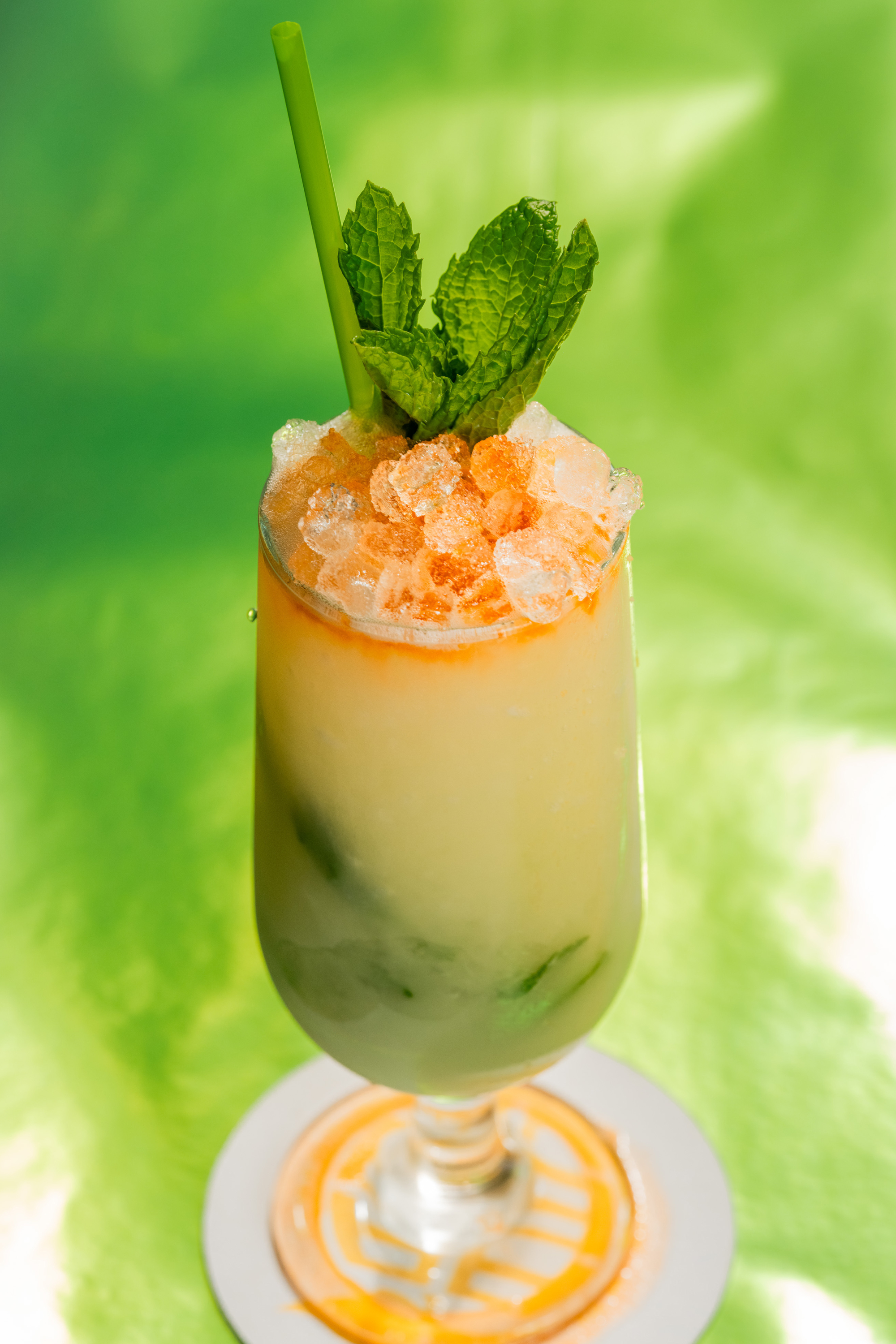 A tall glass with a creamy drink, topped with ice, mint leaves, and a green straw. It sits on a coaster against a vibrant green background.