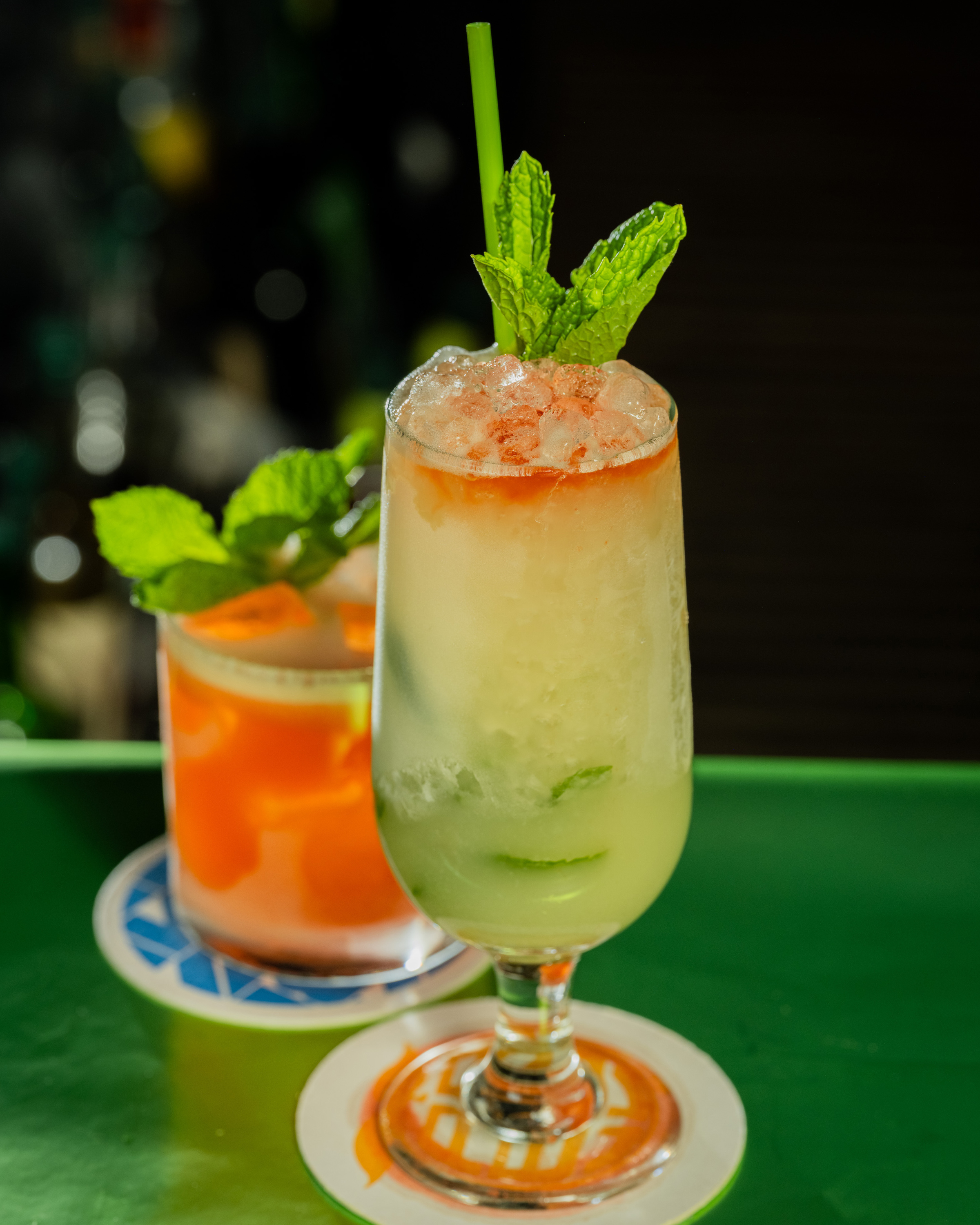 The image shows two cocktails. The front drink is tall, with crushed ice, mint, and a straw; the other is orange, garnished with mint on the side.