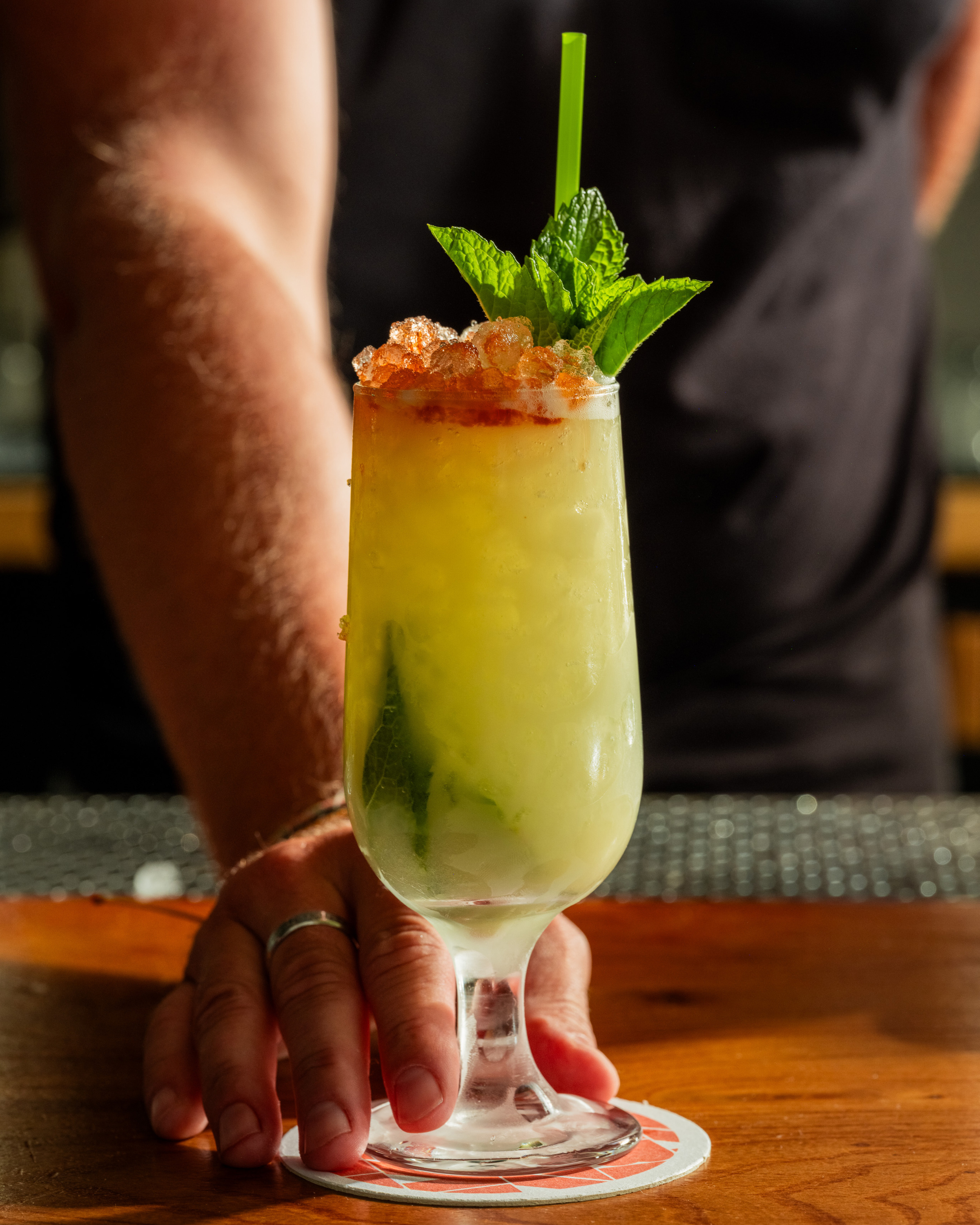 A frosty cocktail in a glass adorned with crushed ice, mint leaves, and a green straw sits on a coaster, held by a hand.
