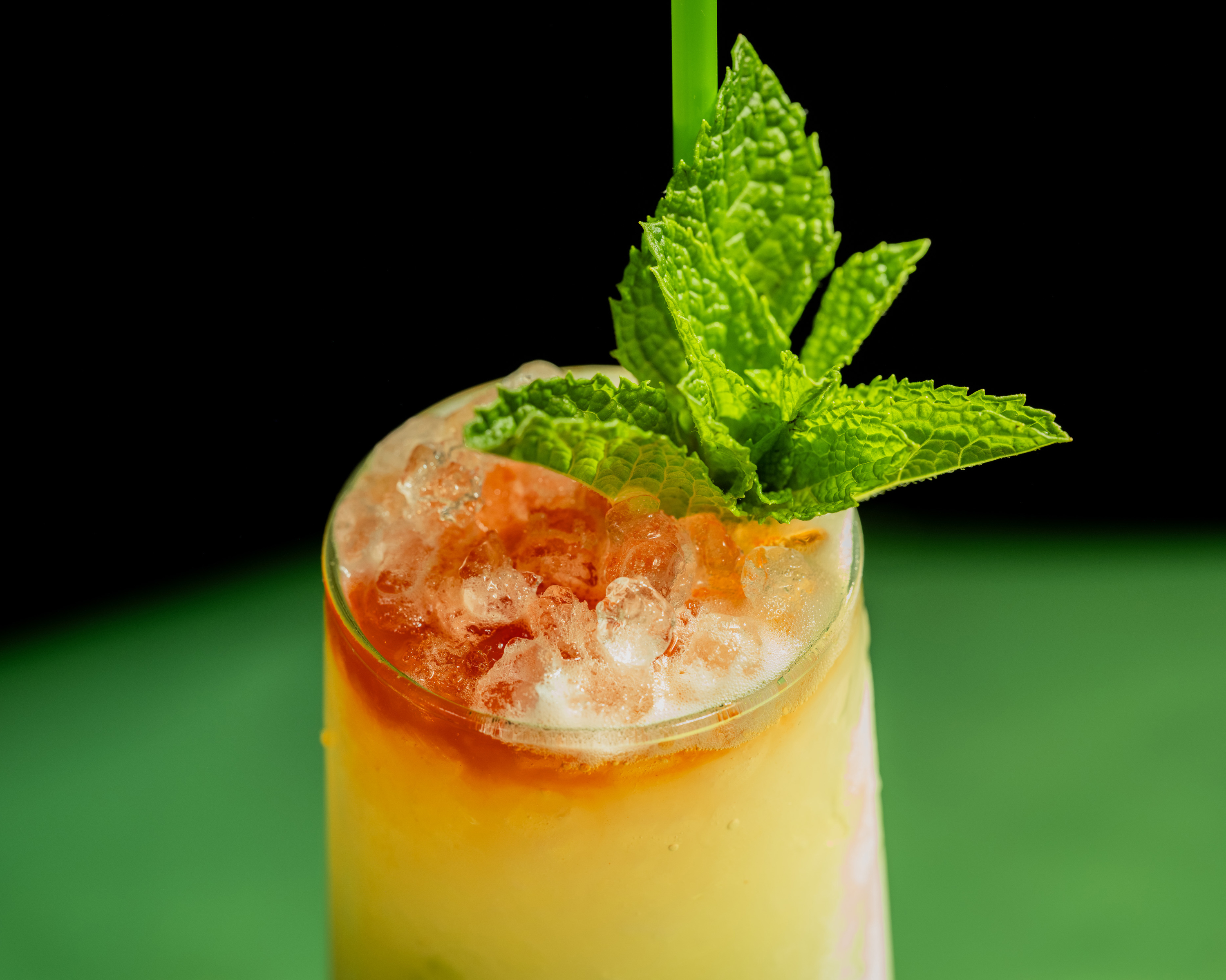 A refreshing drink in a glass topped with crushed ice, vibrant mint leaves, and a green straw; the liquid transitions from yellow to orange.