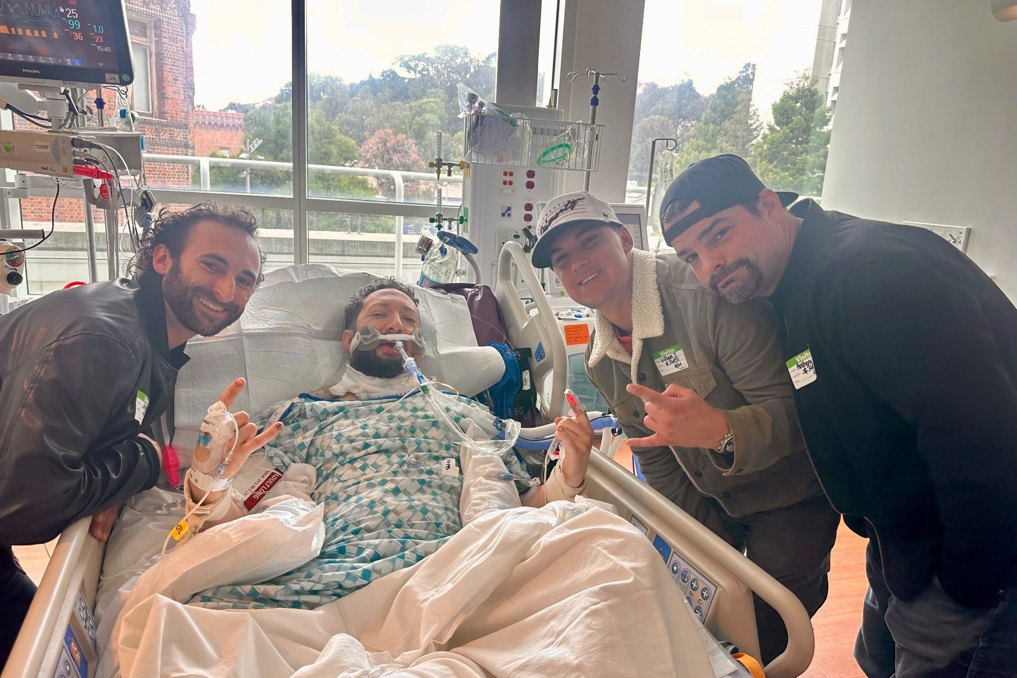 A person is lying in a hospital bed with medical equipment, surrounded by three smiling visitors making hand gestures. There is a window showing greenery outside.