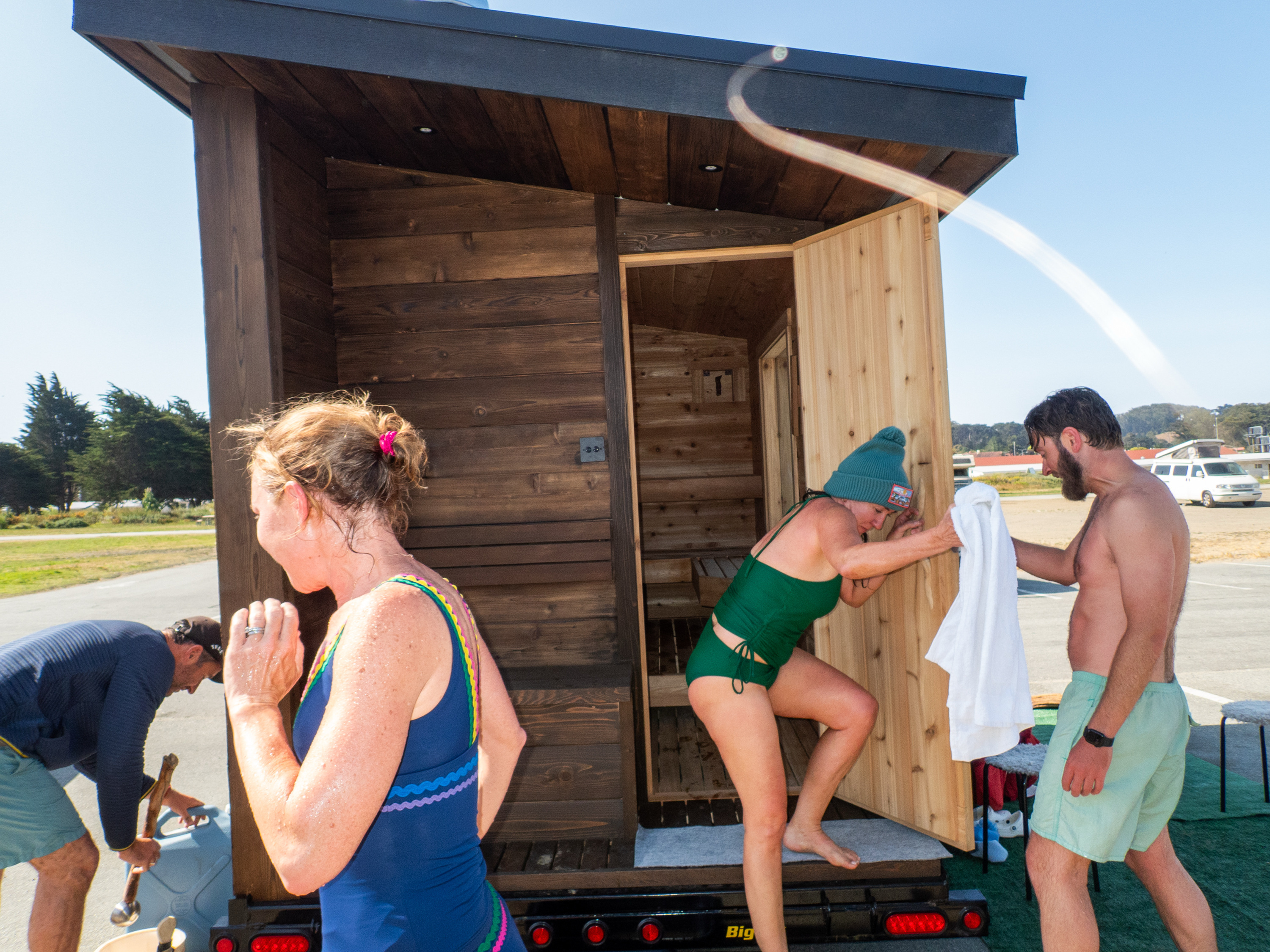 Three people wearing swimwear are outside a small wooden sauna. One person steps out, a towel is held by another, and a third person interacts with a jug.