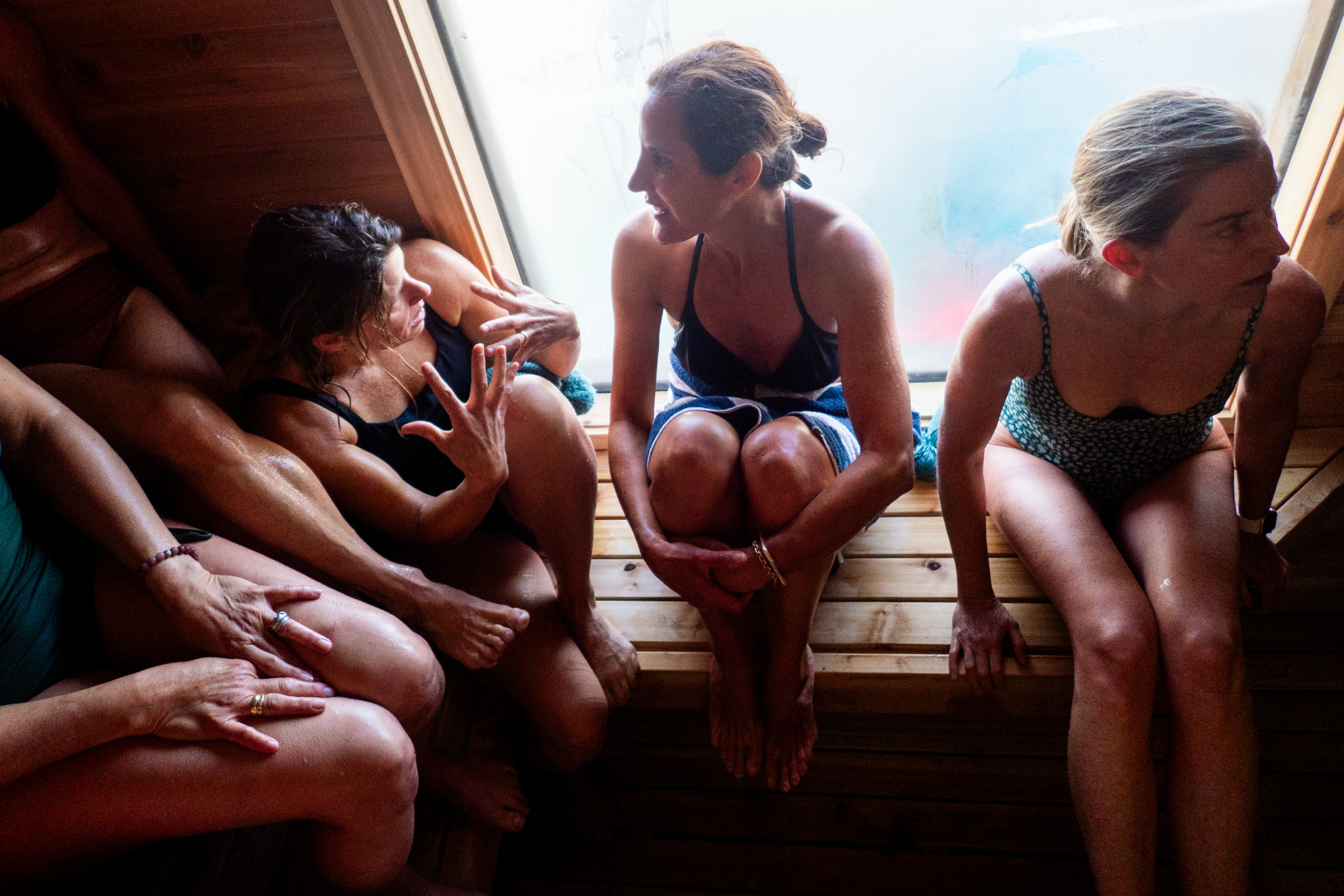 Three women in swimsuits sit closely together on wooden benches in a sauna, engaging in conversation, near a fogged window with light streaming in.