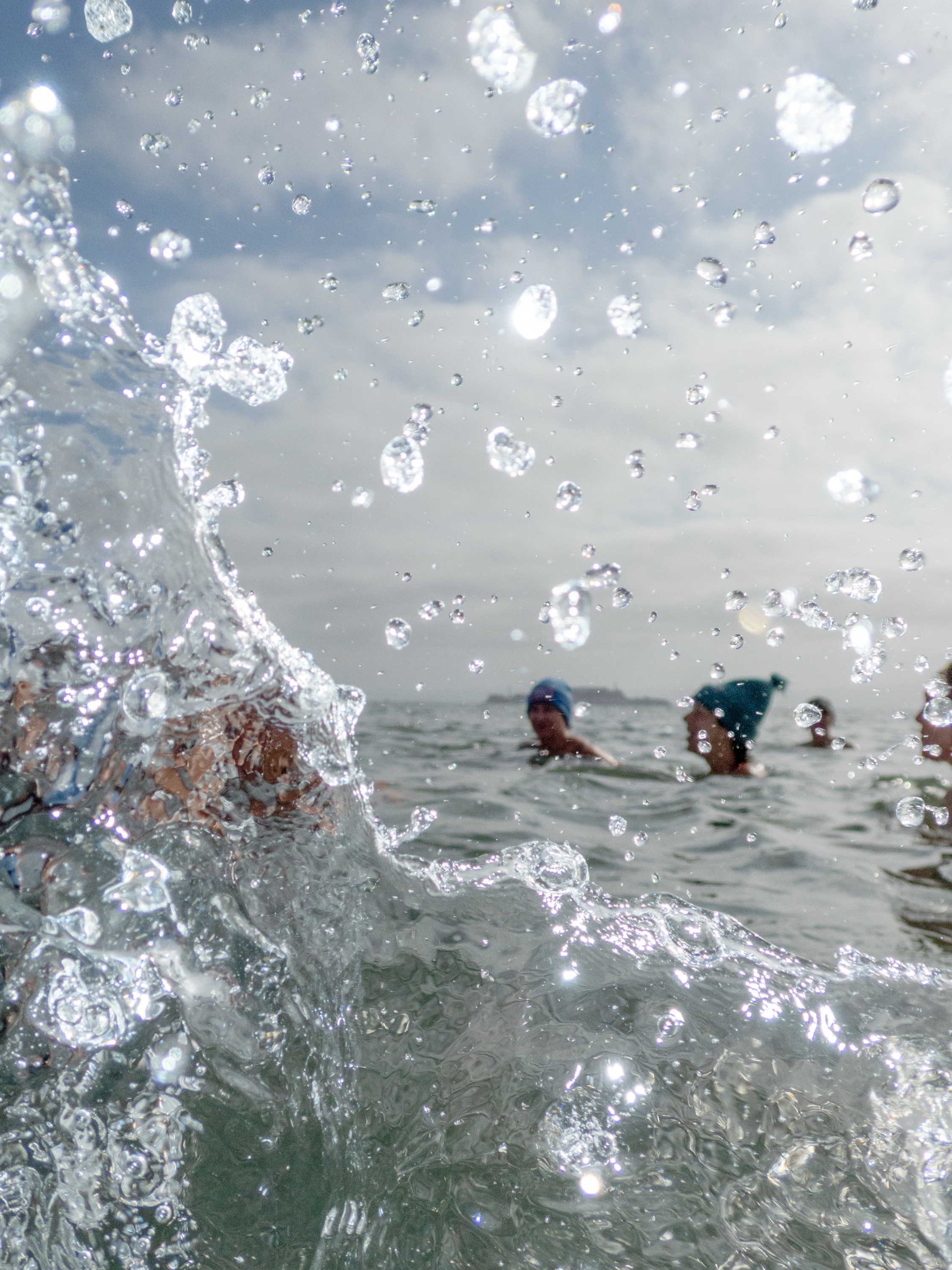 The image shows water splashing in the foreground, with a group of people swimming in the ocean. They wear swim caps, and the sky is cloudy.