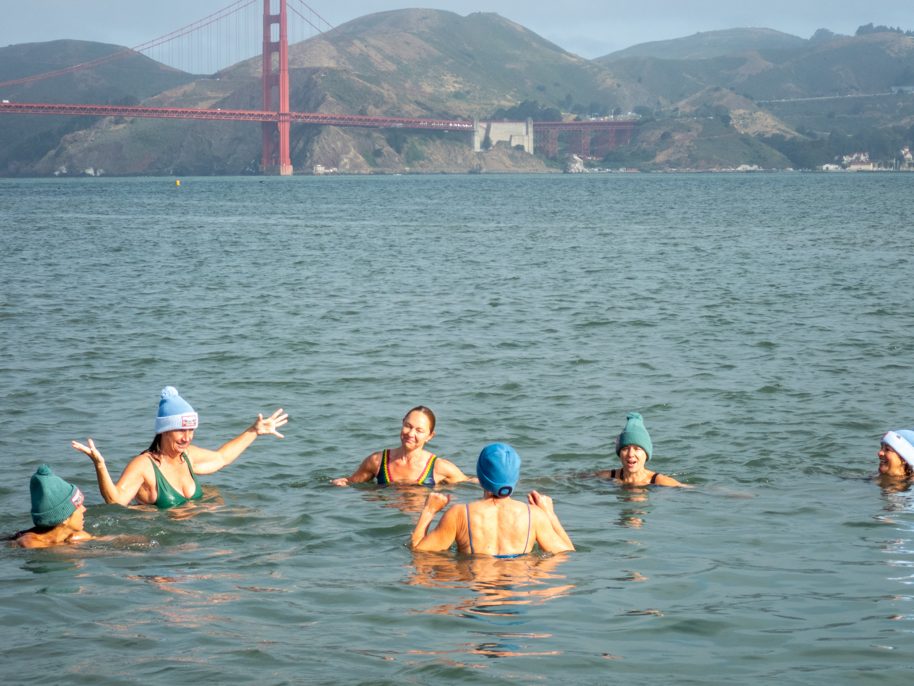A group of people swim in a bay, wearing colorful caps and swimwear. In the background, there's a hilly landscape with a large red bridge spanning across.