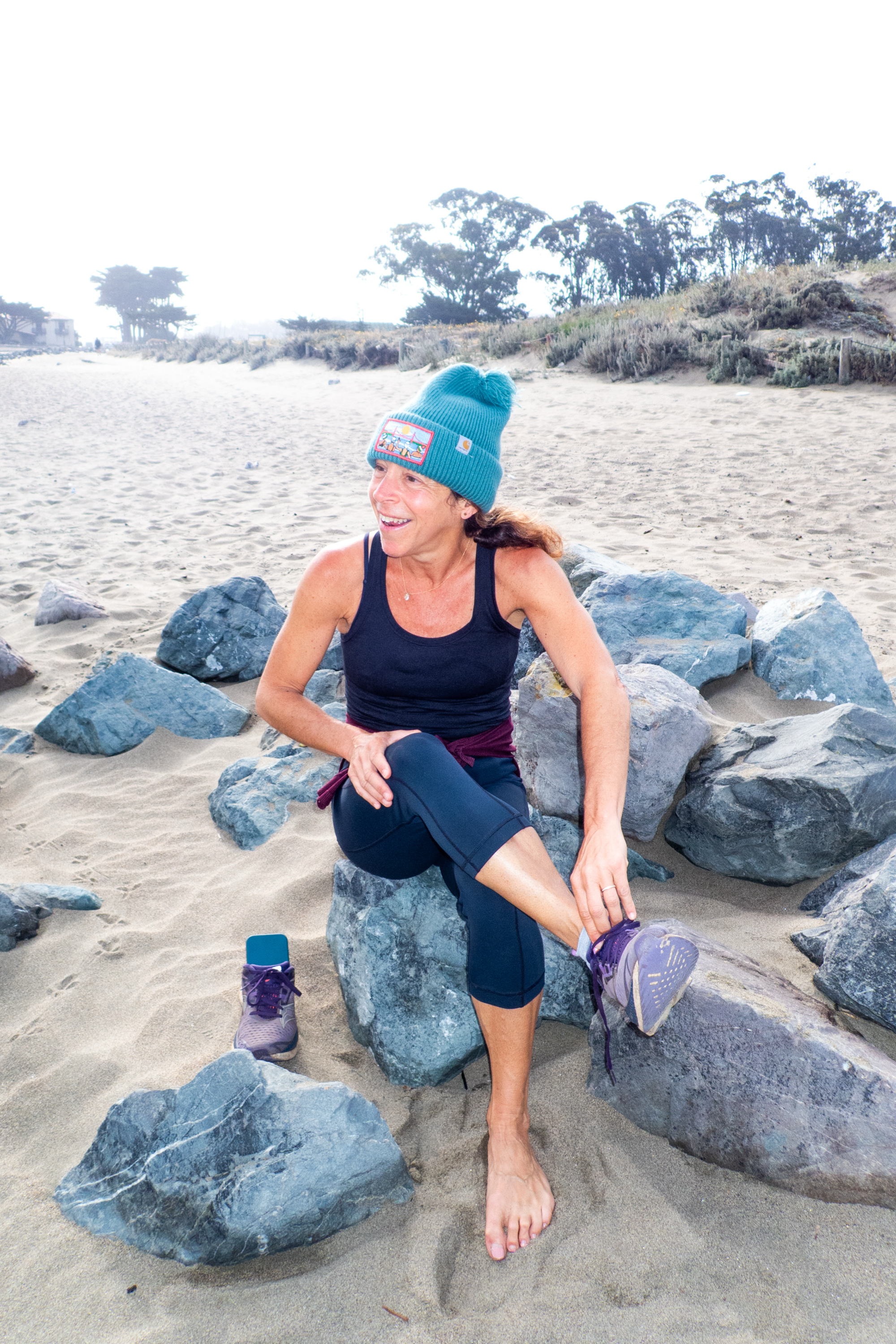 A woman in a turquoise beanie and black tank top sits on rocks at a beach, smiling as she ties her shoe. One shoe is off, and the beach is misty with trees in the background.
