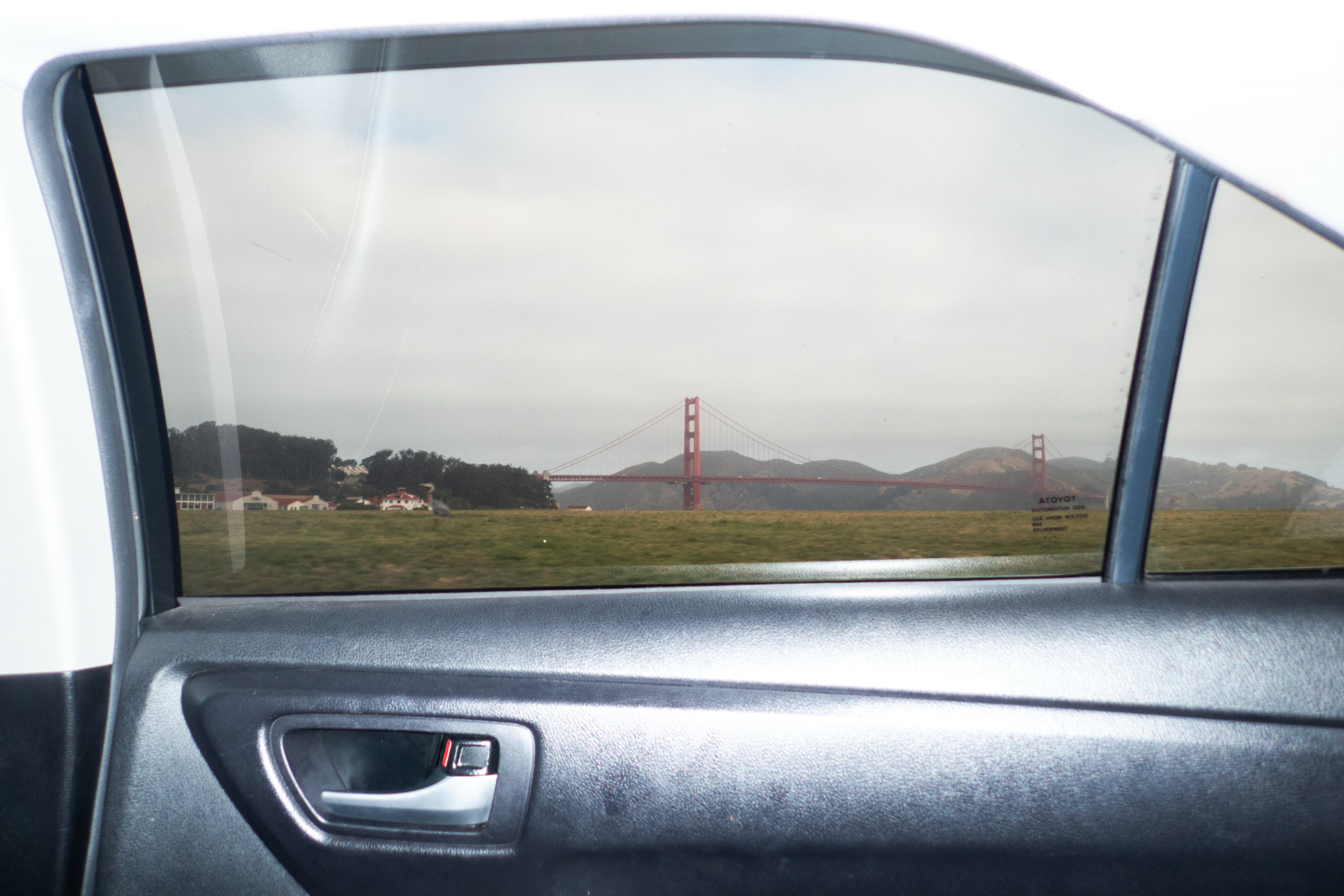 A view of the Golden Gate Bridge is seen through a car window, with green grass and buildings in the foreground under a cloudy sky.