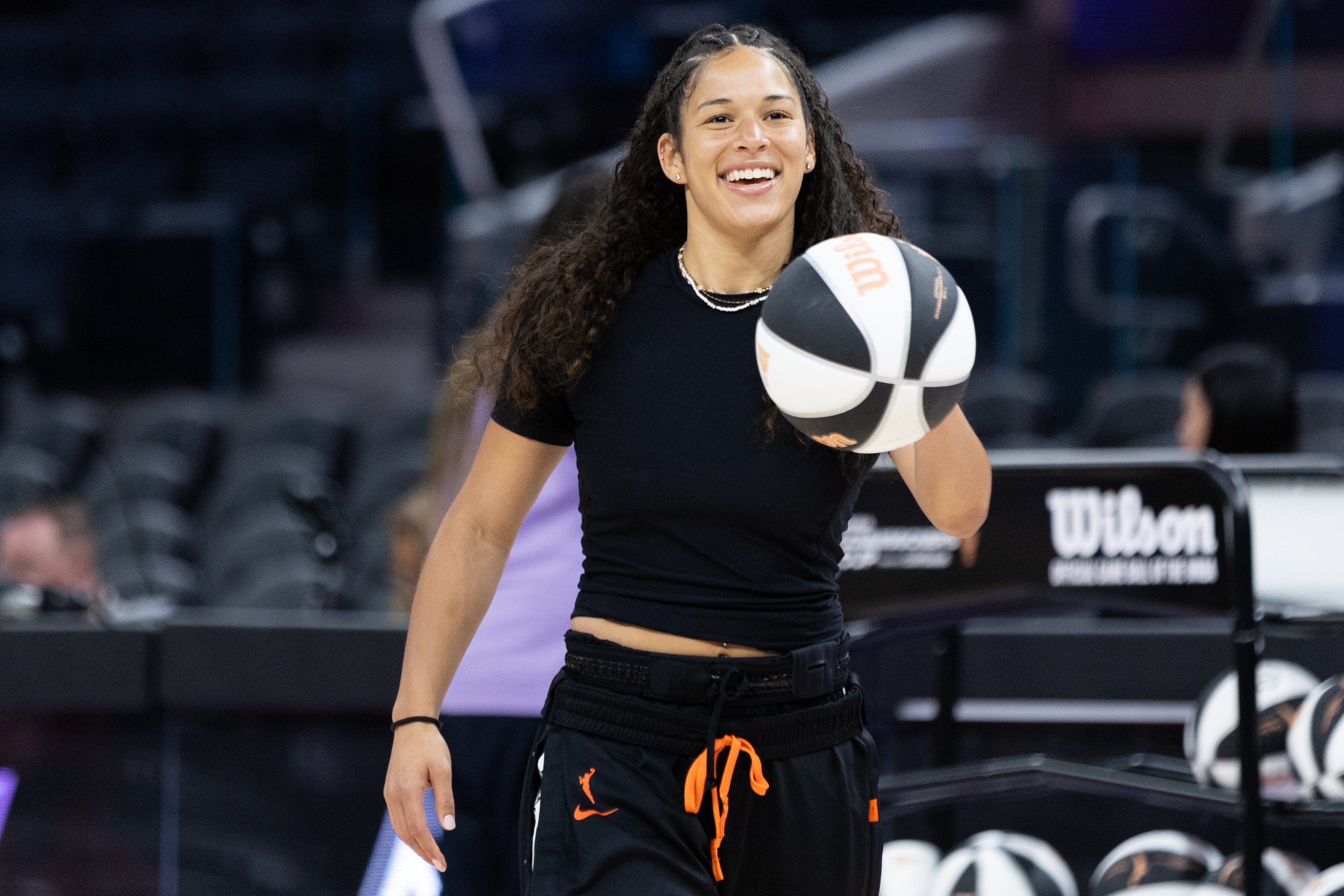 A person with long curly hair smiles while holding a basketball in one hand. They are wearing a black shirt and pants with an orange drawstring.