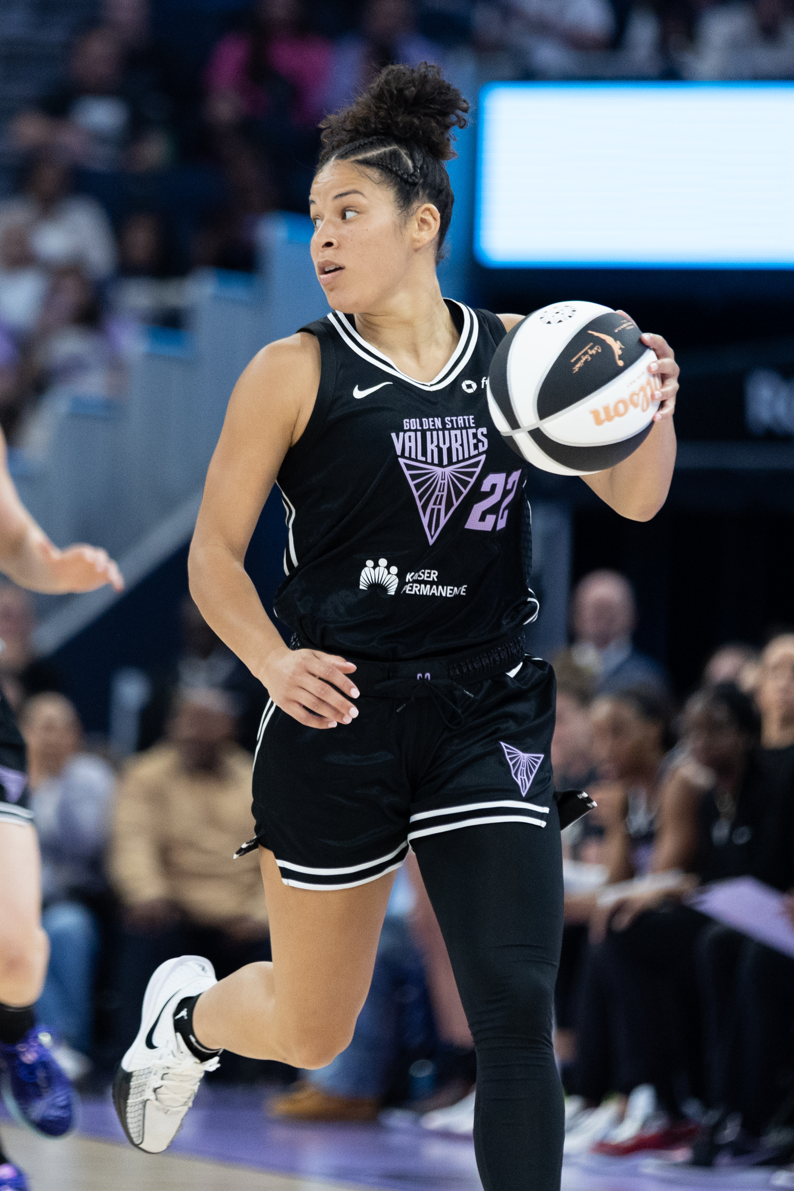 A basketball player in a black &quot;Golden State Valkyries&quot; uniform holds a ball while moving, focused and poised. The background shows blurred spectators.