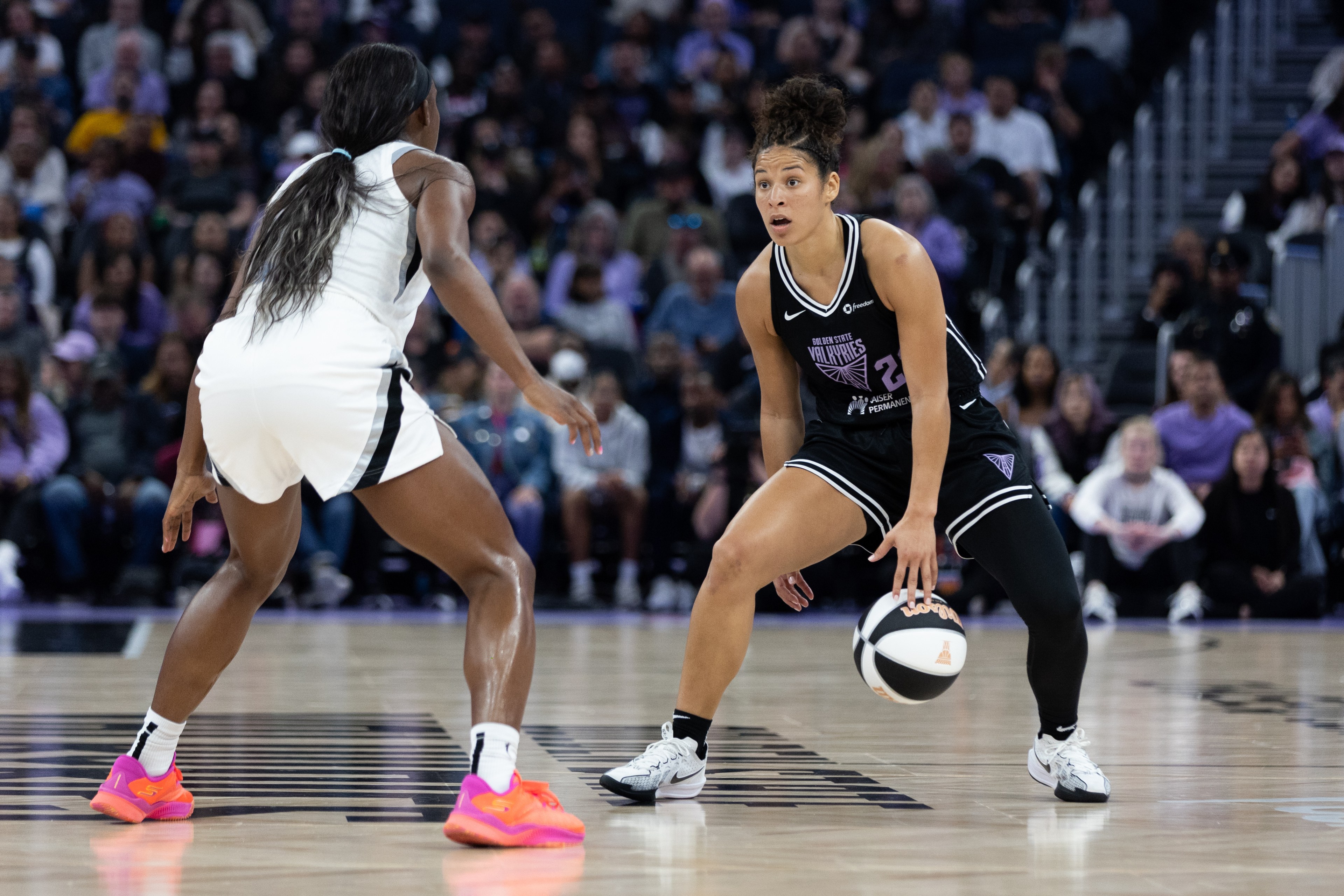 Two basketball players face off on a court. One in black dribbles the ball, while the other in white defends. A crowd watches from the stands.