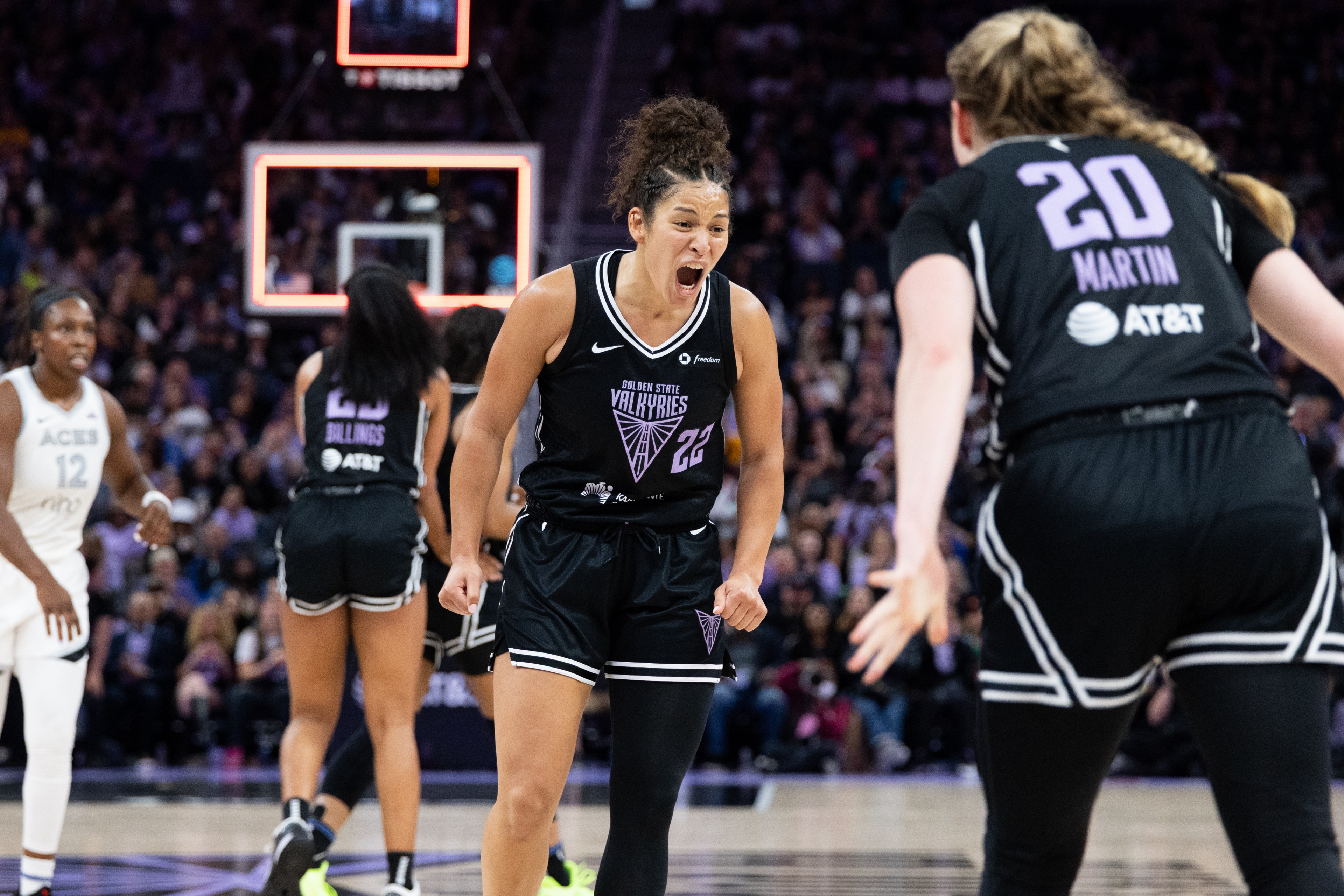 A basketball player in a black jersey with &quot;Golden State Valkyries&quot; shouts on the court, facing another player. The background shows a crowded arena.