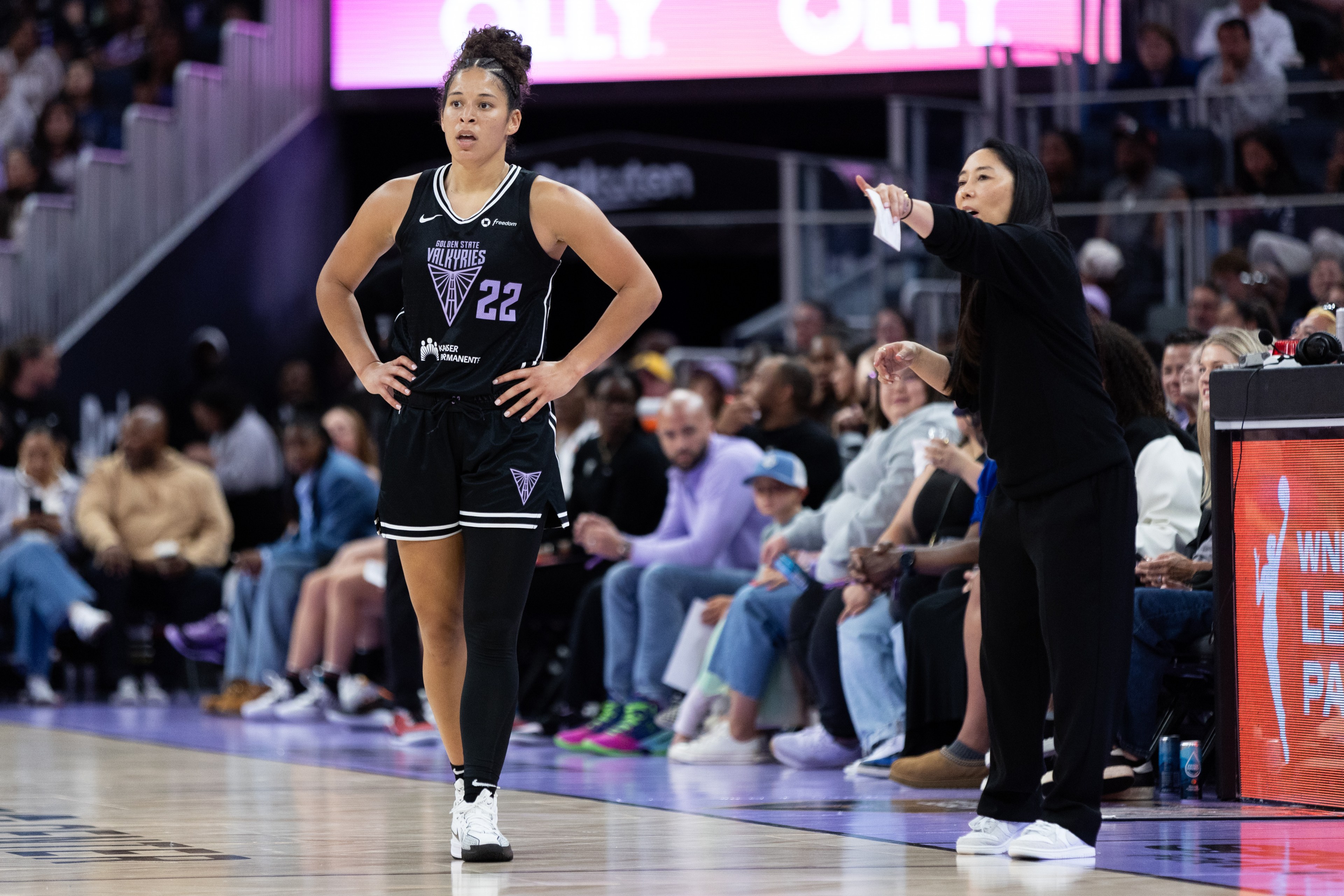 A basketball player stands on the court in a black uniform with &quot;22&quot; on it, while a coach gestures and speaks from the sidelines. Spectators watch in the background.