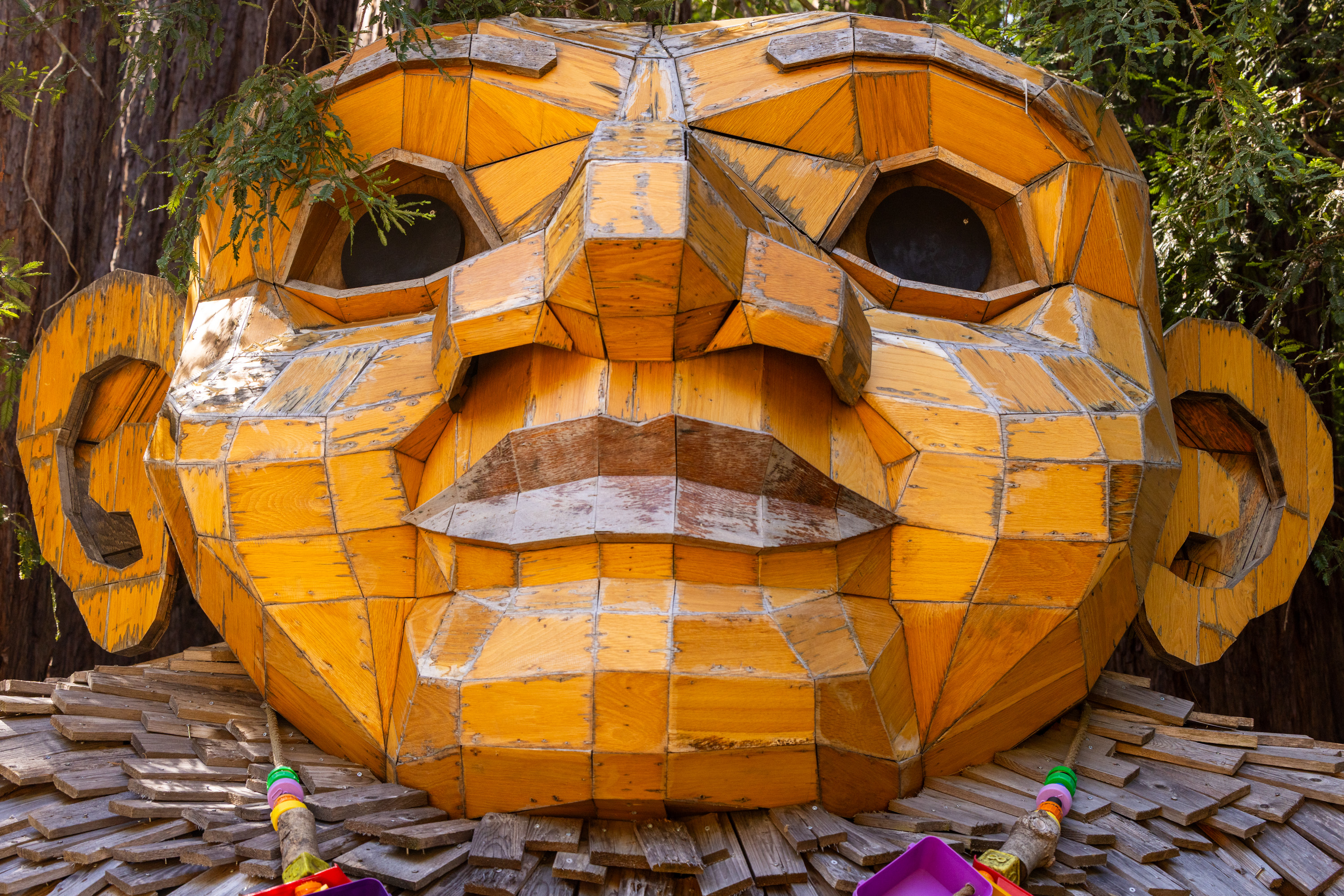 The image shows a large, geometric wooden sculpture of a face with prominent features. It has big ears and is constructed from yellowish wood panels.