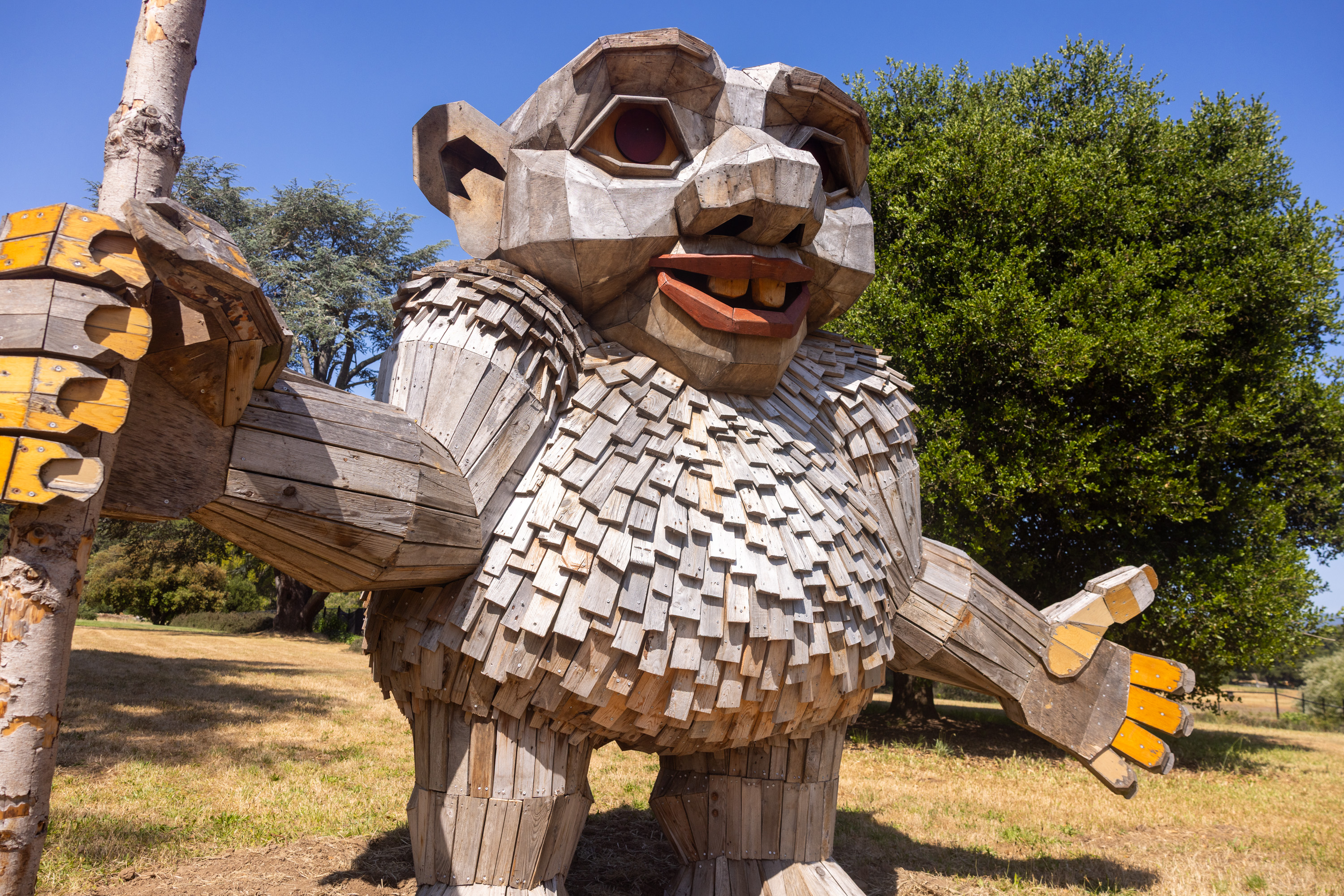 A large, whimsical wooden troll sculpture stands outdoors, made from various wooden planks. It holds a tree trunk and has a cheerful expression.