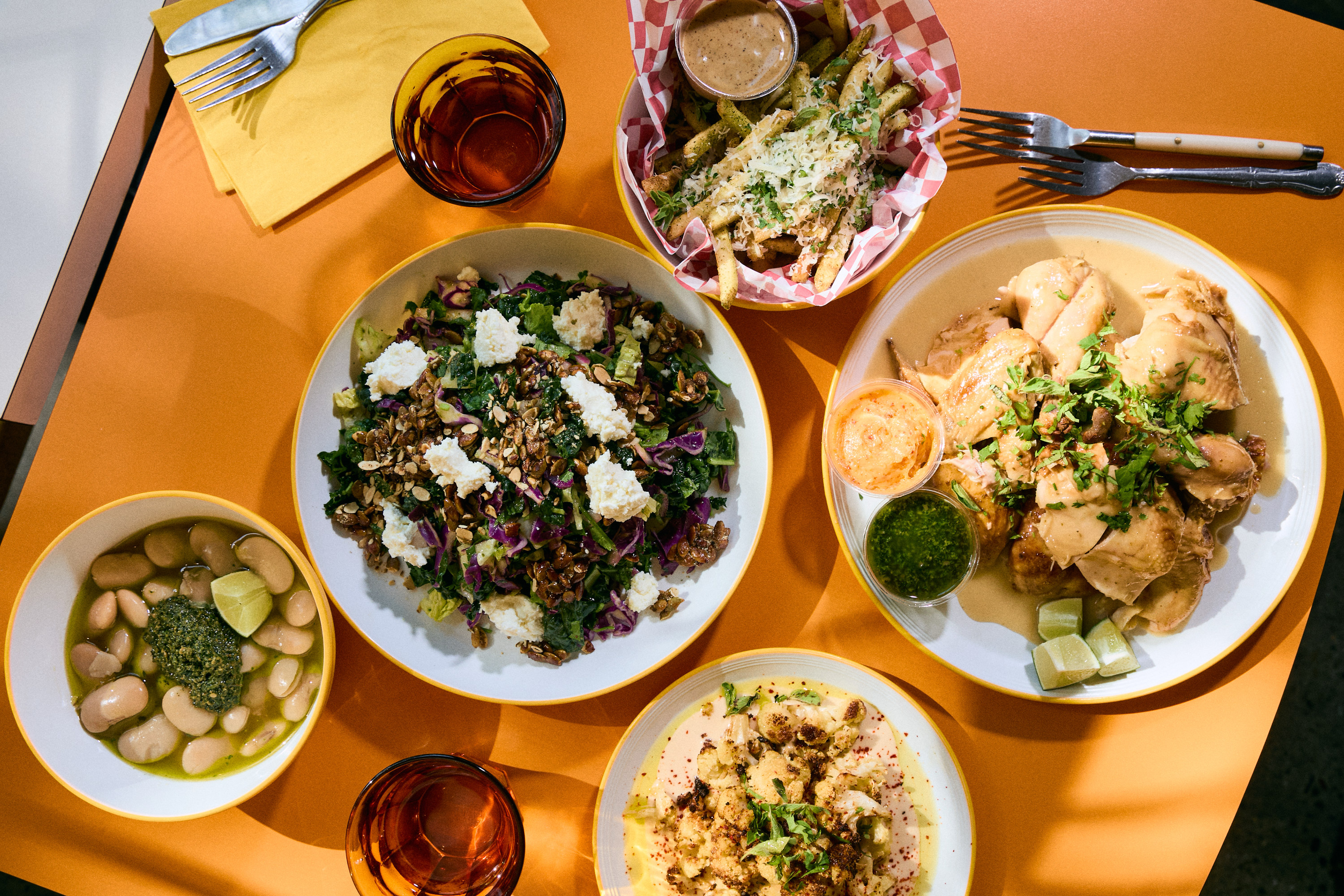 A vibrant table with roasted chicken, colorful salad, fries with sauce, beans, and two drinks on an orange surface with cutlery and napkins.