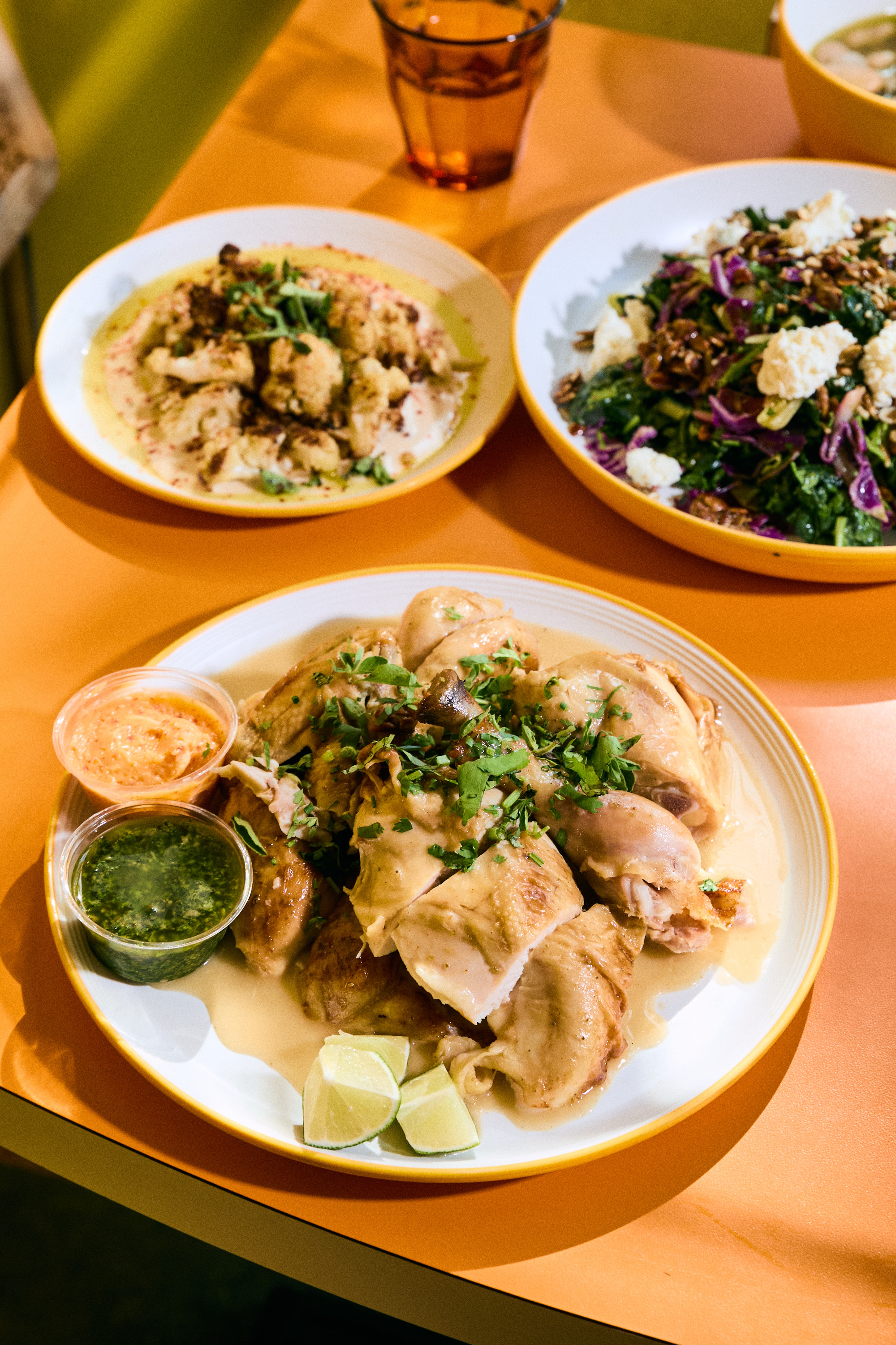 A plate of roasted chicken with herbs, lime wedges, and two dipping sauces is foregrounded. In the background are bowls of cauliflower and a mixed salad.