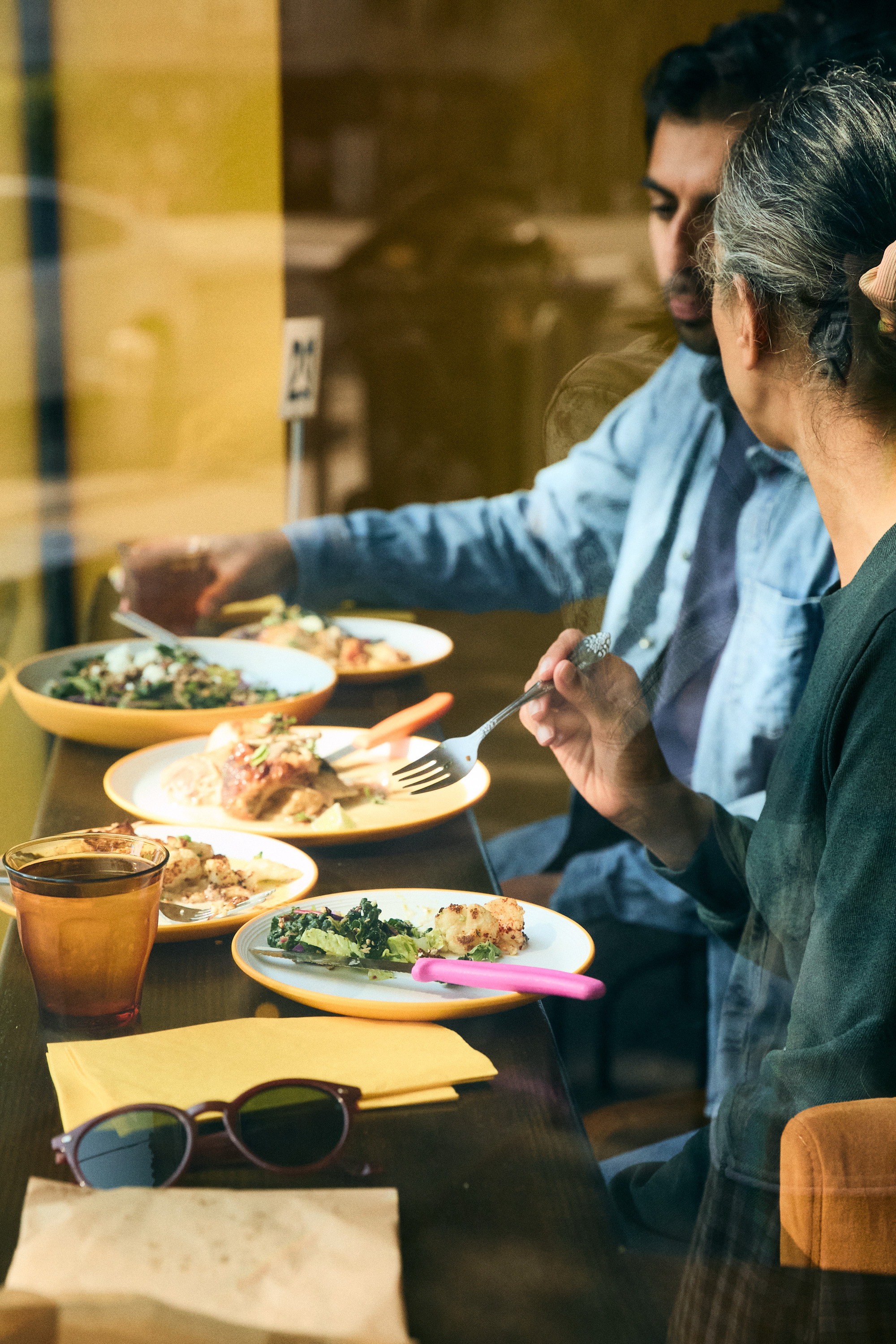 Two people dine by a window with plates of food, a fork, drinks, and sunglasses on the table, in warm lighting.