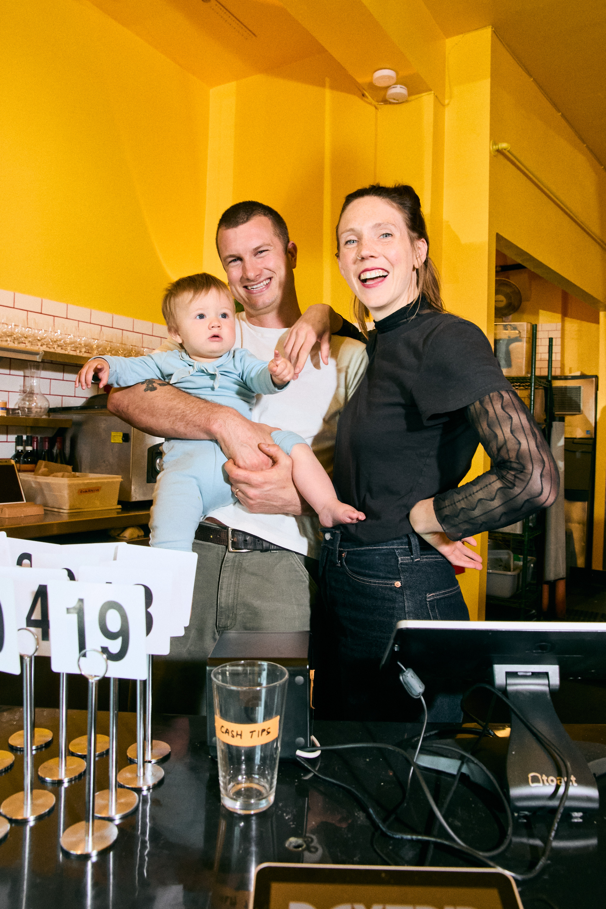 A smiling couple stands in a bright yellow room, holding a baby. The baby is wearing a blue outfit. Nearby, there are number signs and a tip jar.