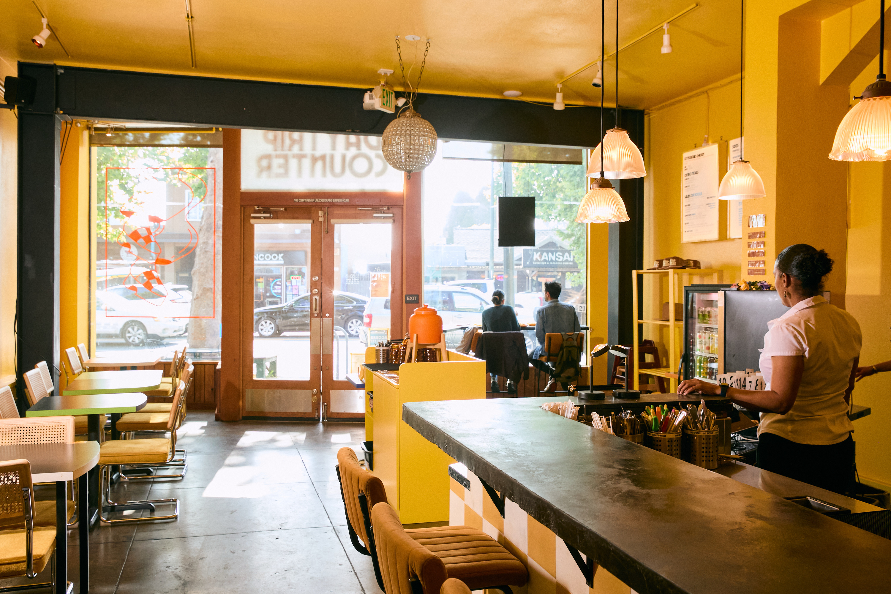 A cozy cafe with yellow walls, tables, and chairs, a counter with utensils, hanging lights above, and two people sitting by the window.