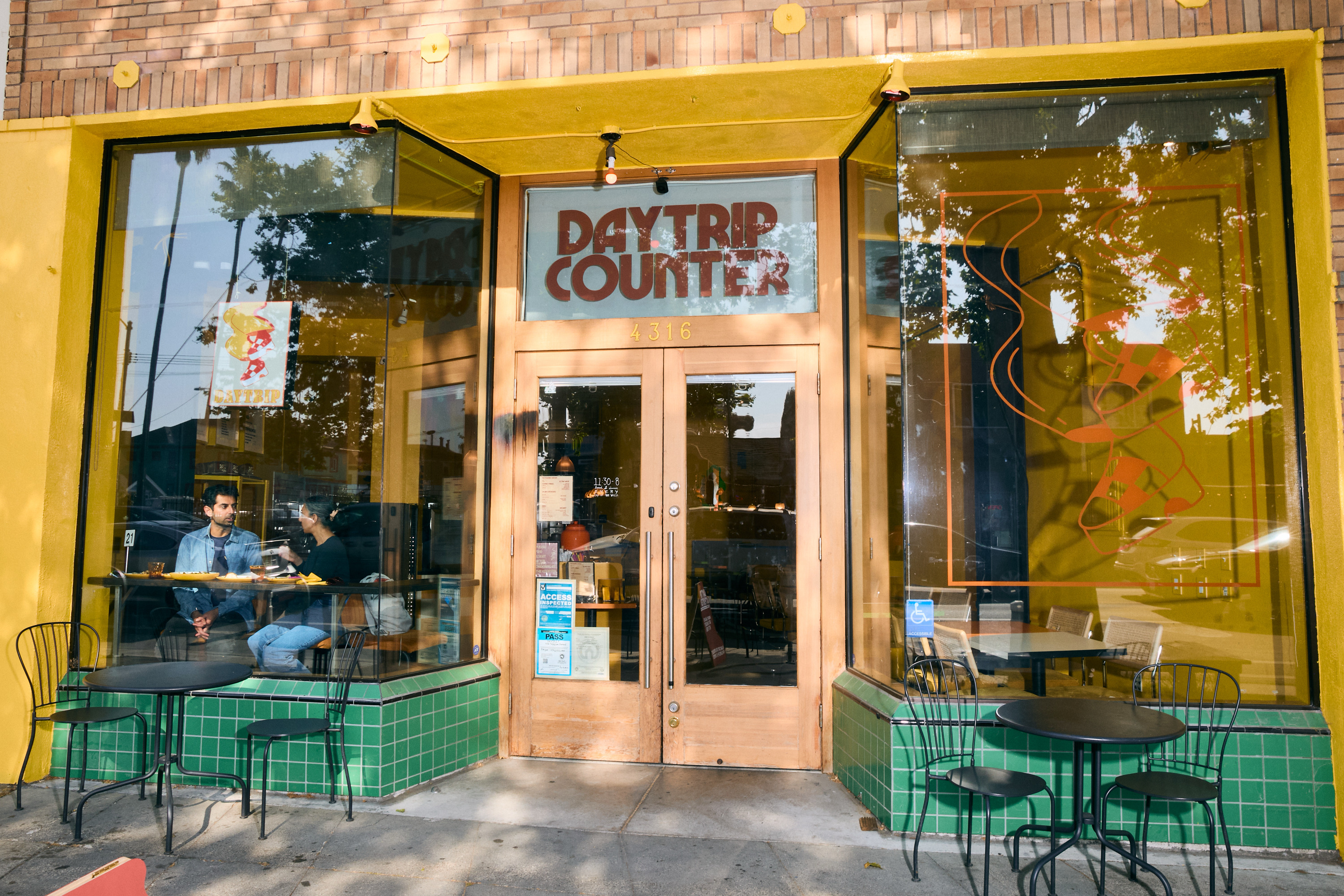 The image shows a cafe named &quot;Daytrip Counter&quot; with a glass facade. Two people sit inside at a table, and there are empty tables and chairs outside.