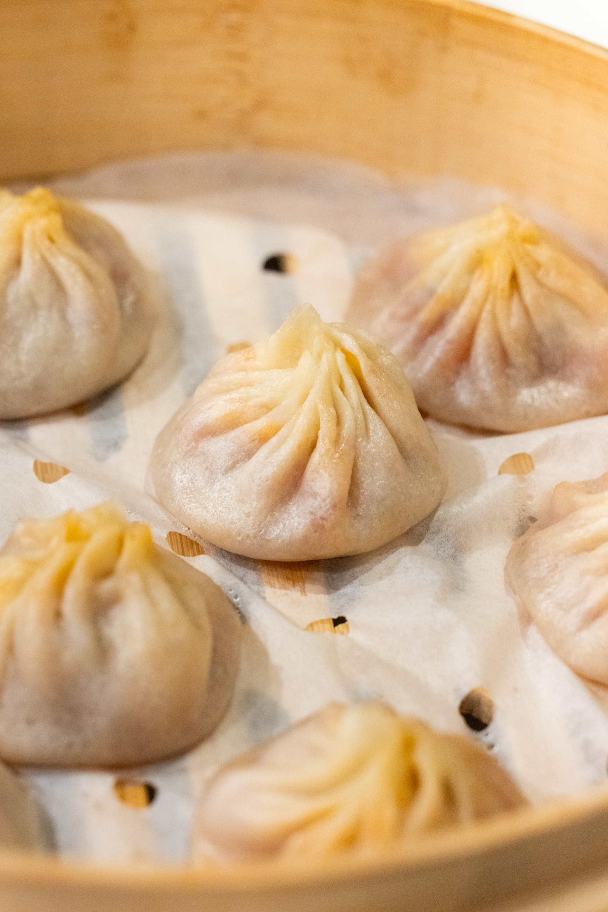 The image shows a bamboo steamer with five plump, steamed dumplings, slightly glossy, with pleated tops on parchment paper.