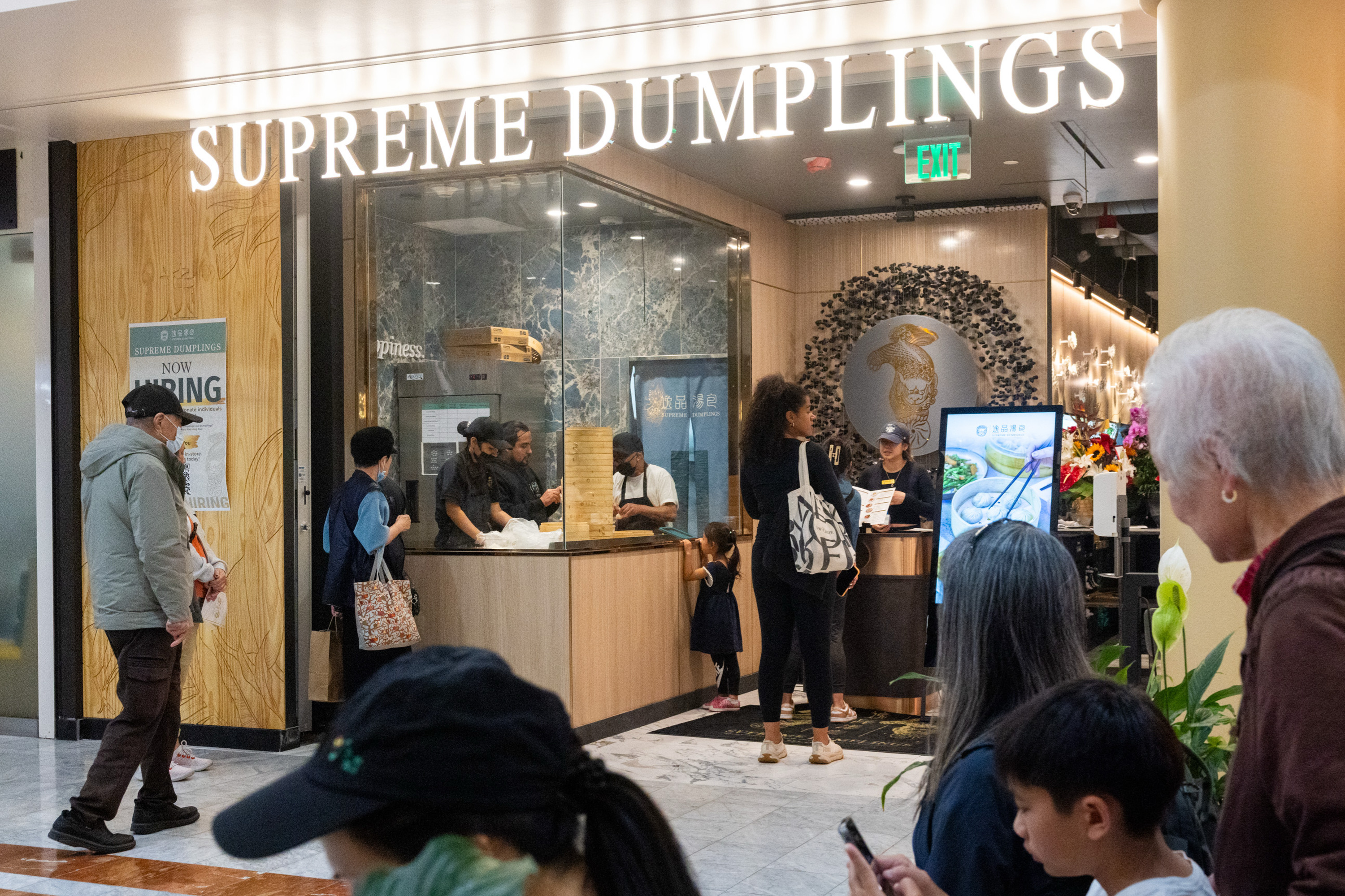 The image shows a dumpling restaurant with a sign reading "Supreme Dumplings." Several people are waiting in line, and staff members are preparing food inside.