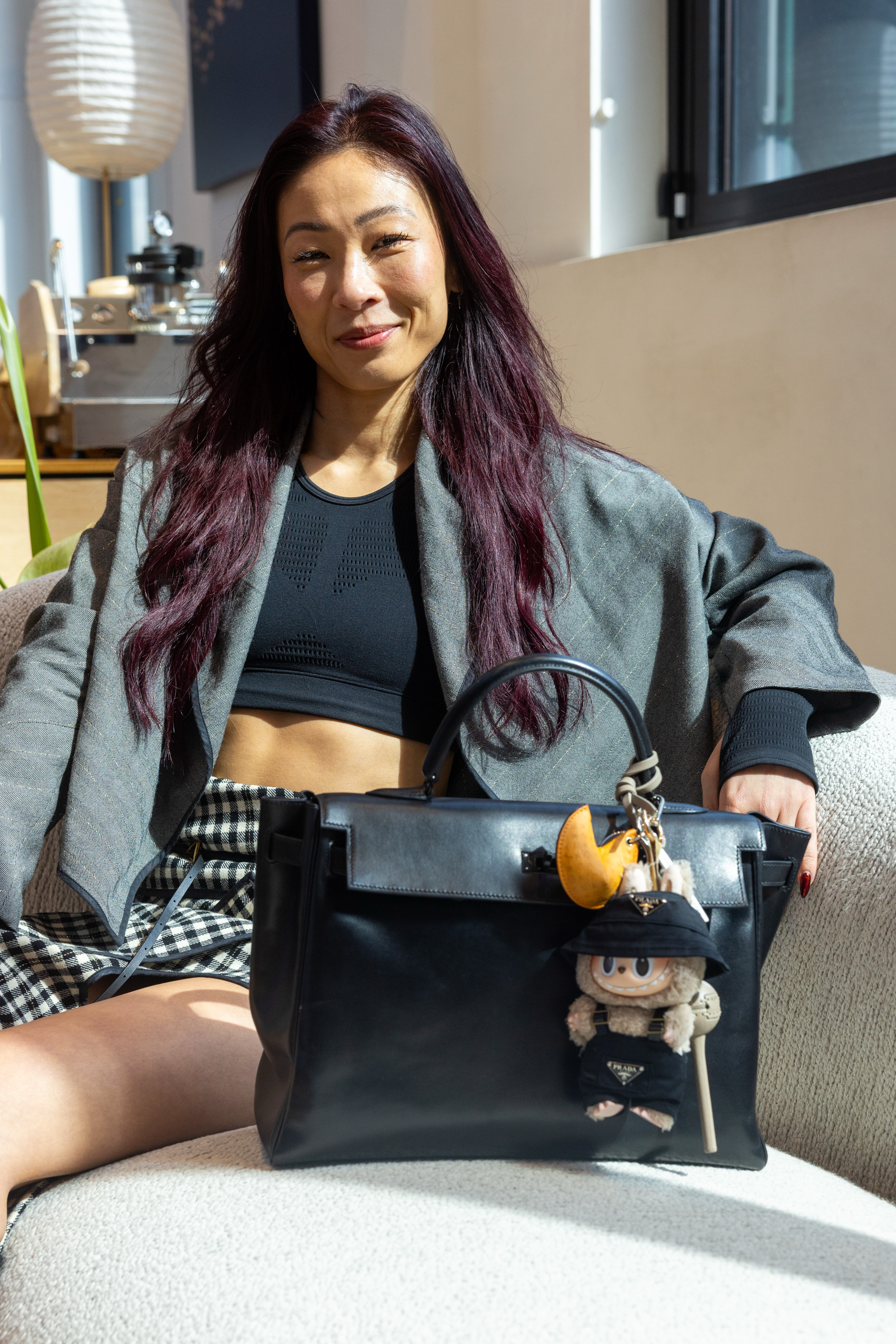 A person with long, dark red hair sits on a chair. They're wearing a black top, gray blazer, and checkered skirt. A black handbag with a doll charm is beside them.