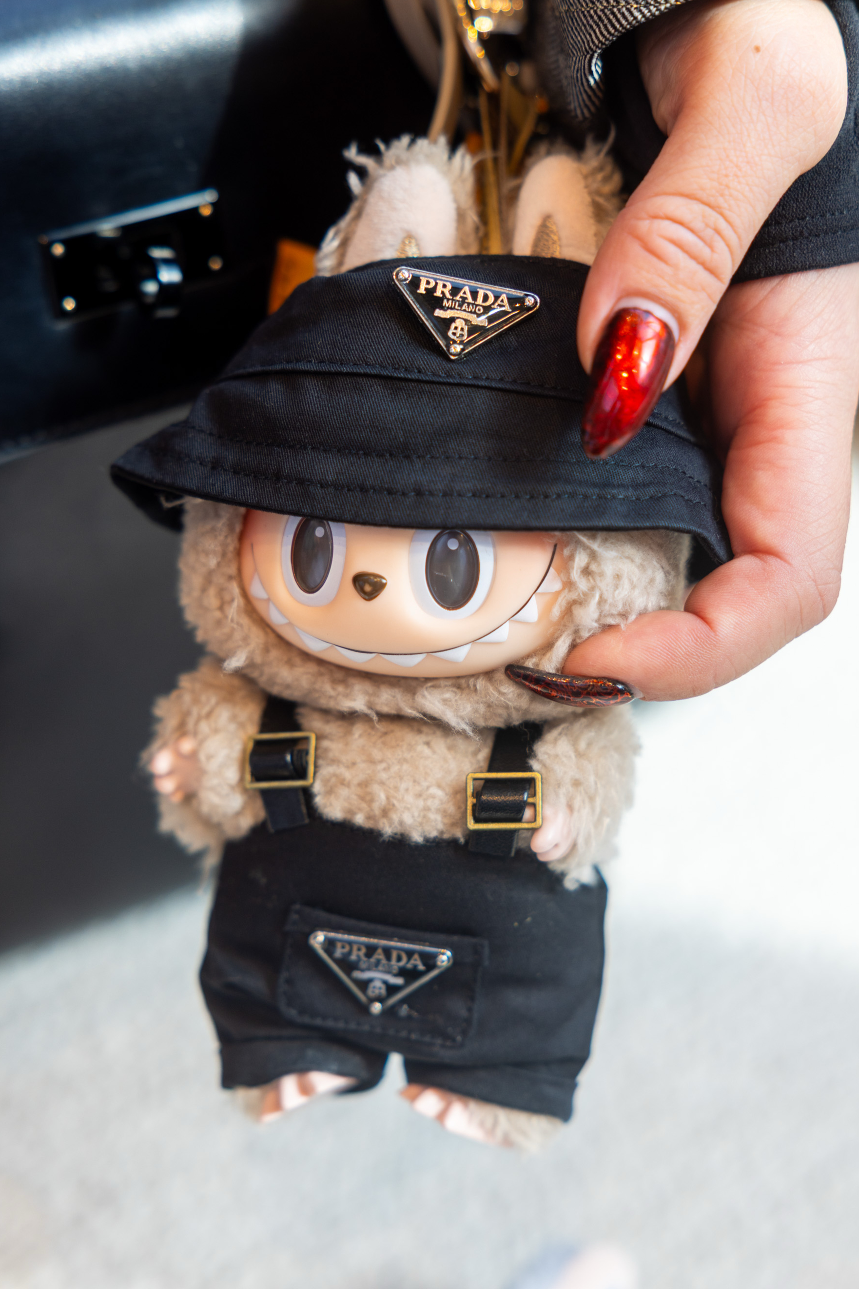 A plush doll with large eyes and a wide smile wears a black Prada hat and overalls. A hand with red nails holds it, accentuating the luxury brand.