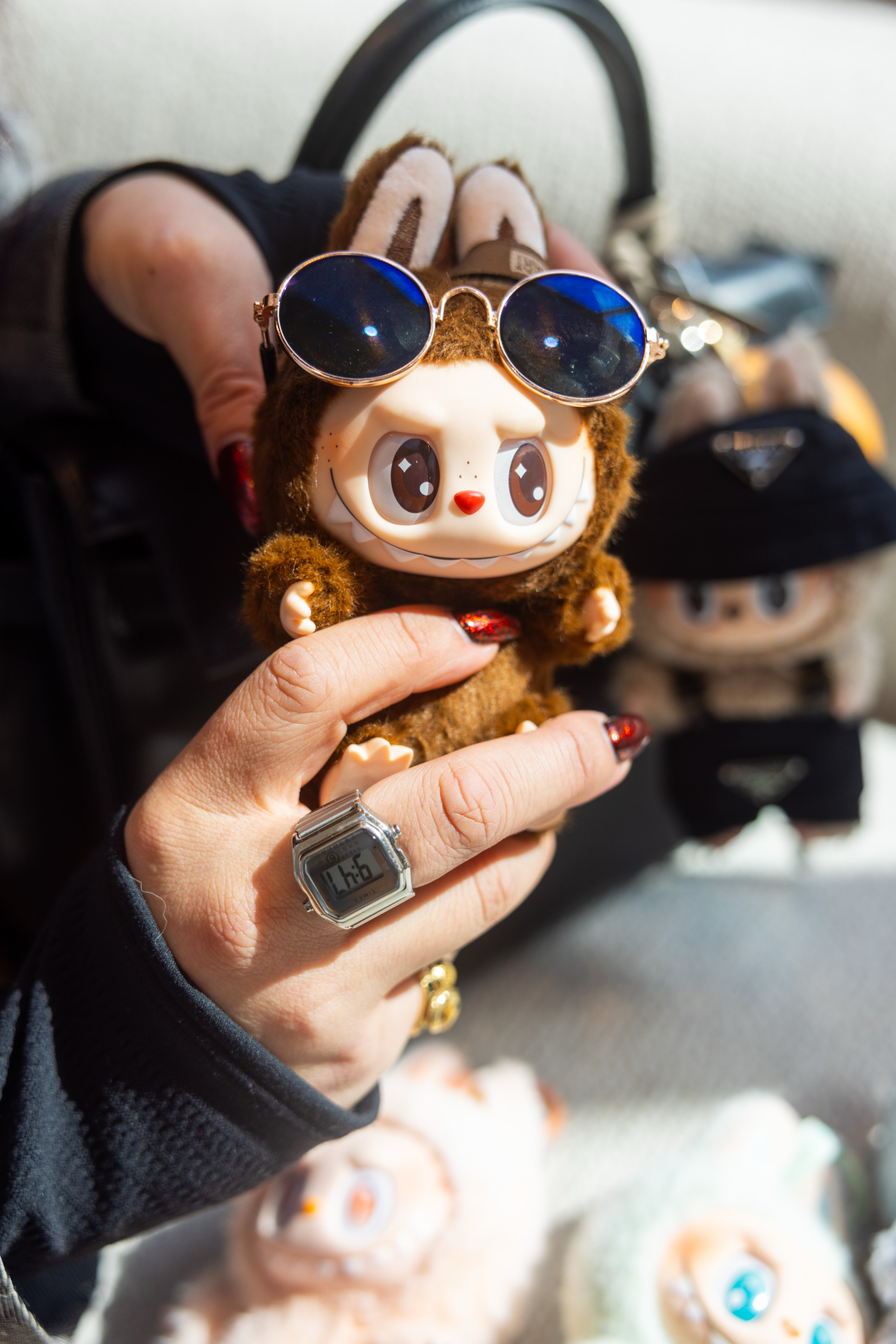 A hand holds a cute toy wearing blue sunglasses, with a digital watch visible on the wrist. Other similar toys are blurred in the background.