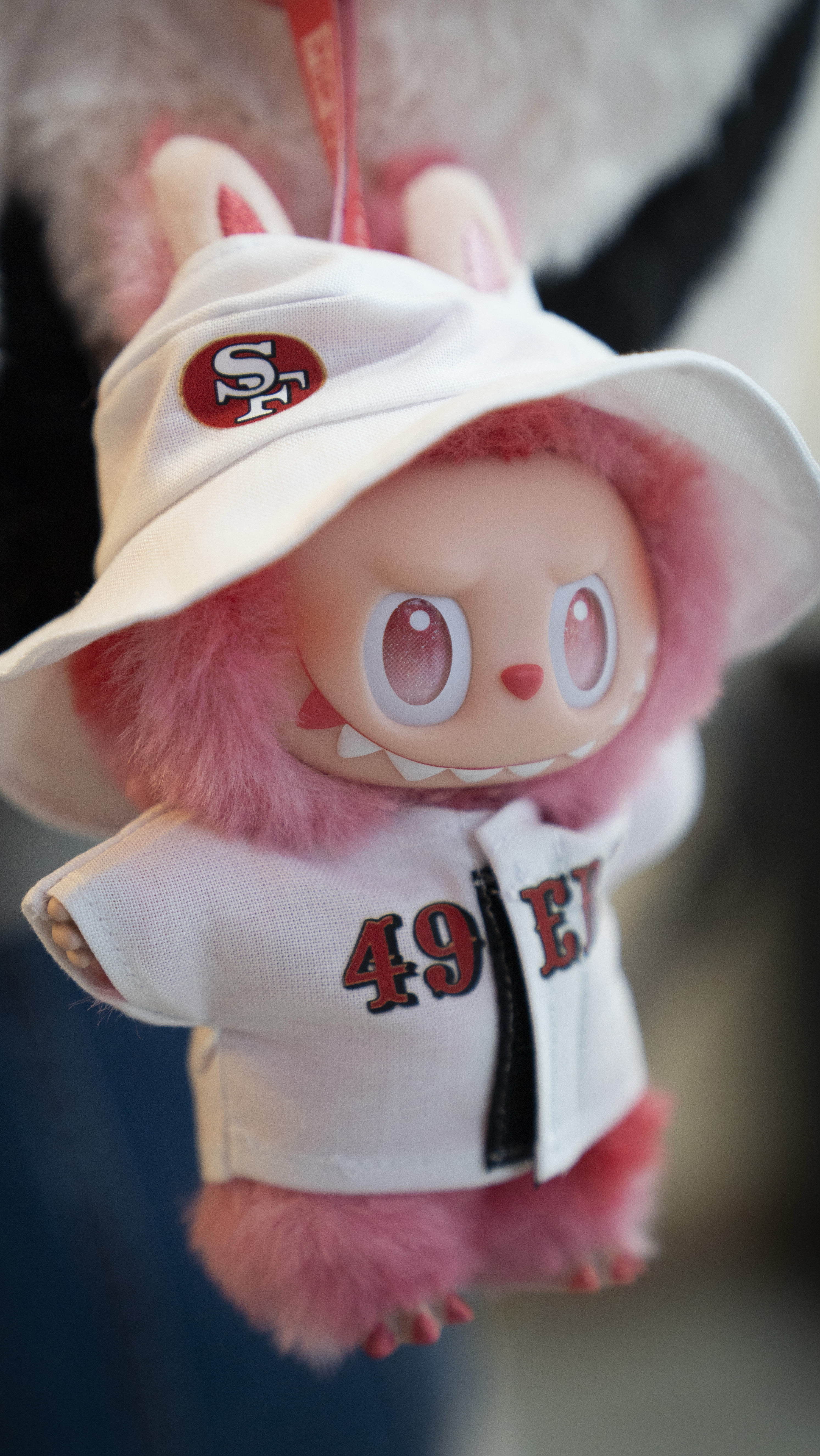A cute pink furry toy with large eyes wears a white jacket with &quot;49ERS&quot; and a matching hat featuring the SF logo.