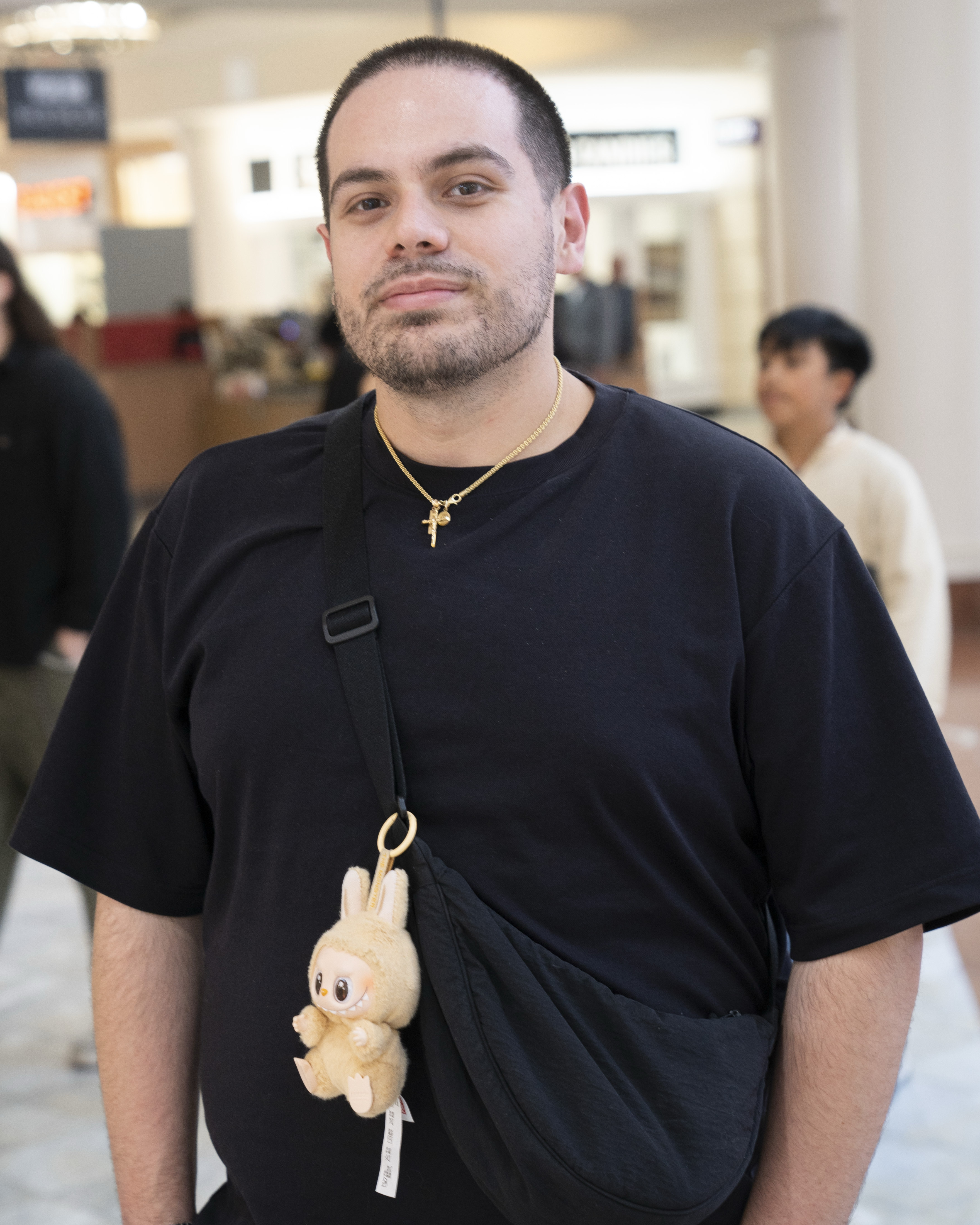 A person with short hair and a beard is wearing a black shirt and cross necklace. They have a black crossbody bag featuring a cute plush toy attached.