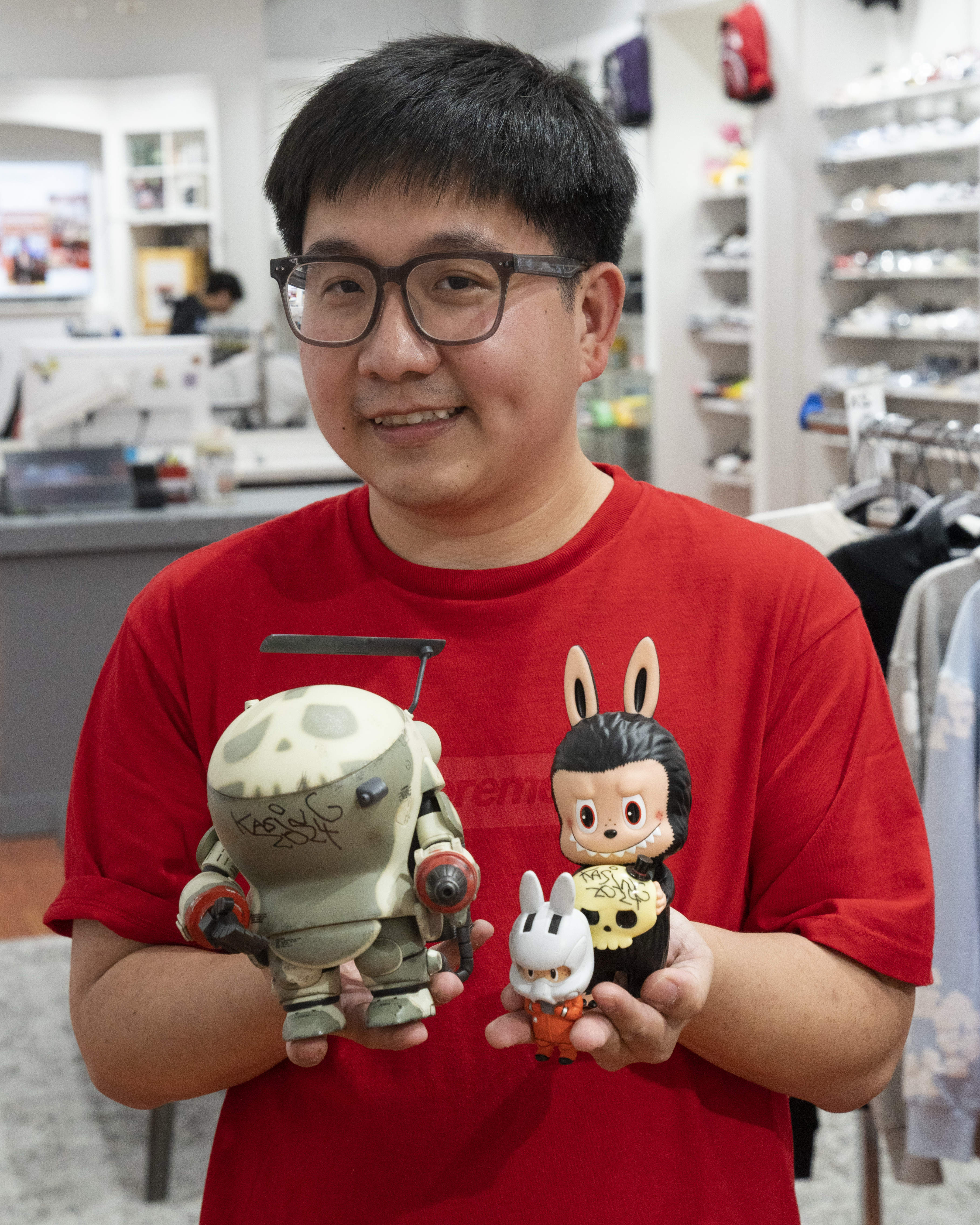 A person in a red shirt and glasses holds three toy figurines: a gray robot with graffiti, a character with rabbit ears, and a small rabbit-hooded figure.