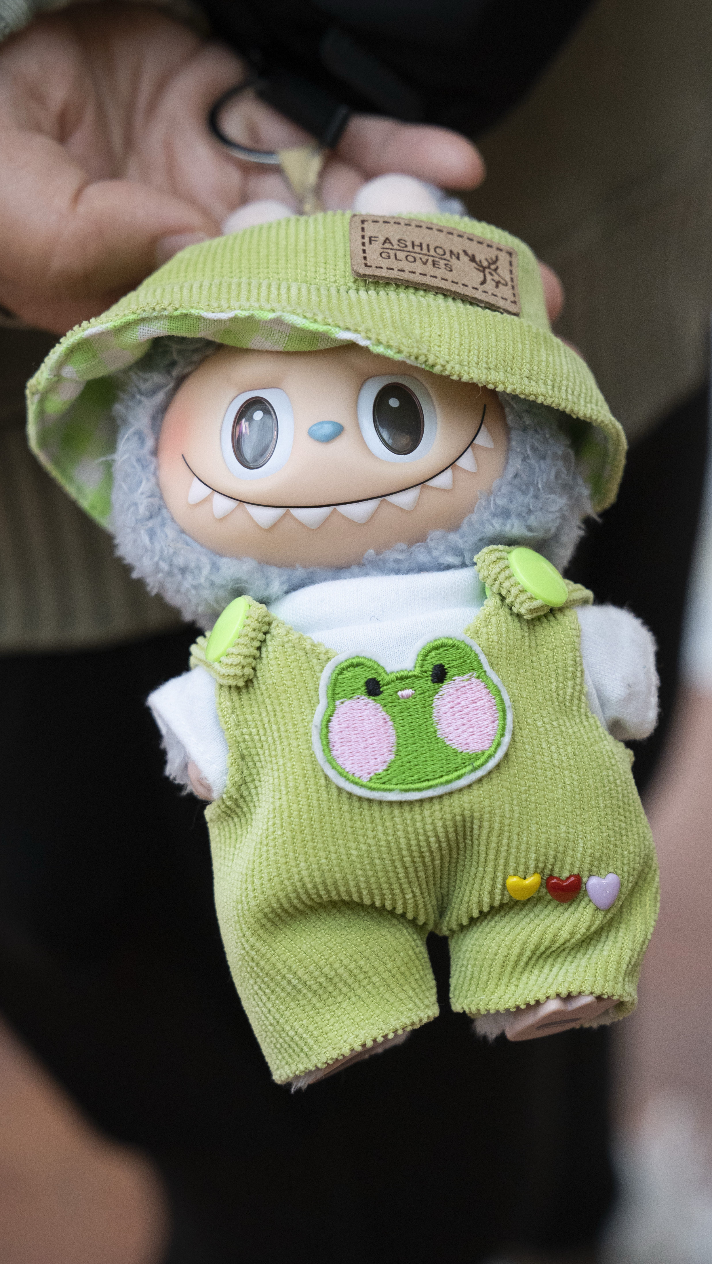A cute doll with a big smile is wearing a green hat and overalls with a frog patch. Three heart buttons are on its leg. A hand is holding the doll.