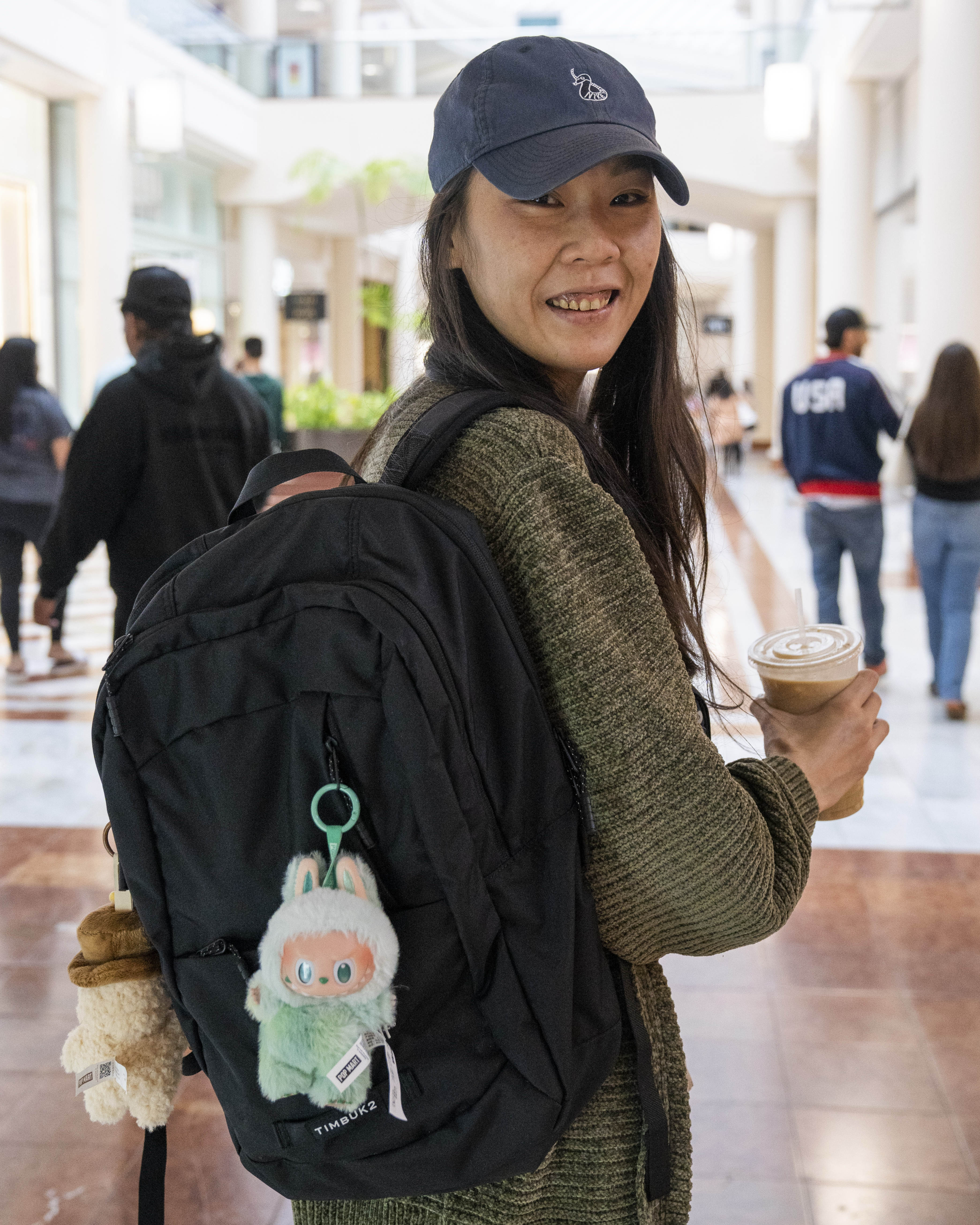 A person wearing a cap and sweater smiles while holding a beverage in a shopping mall. They carry a black backpack adorned with a plush toy keychain.